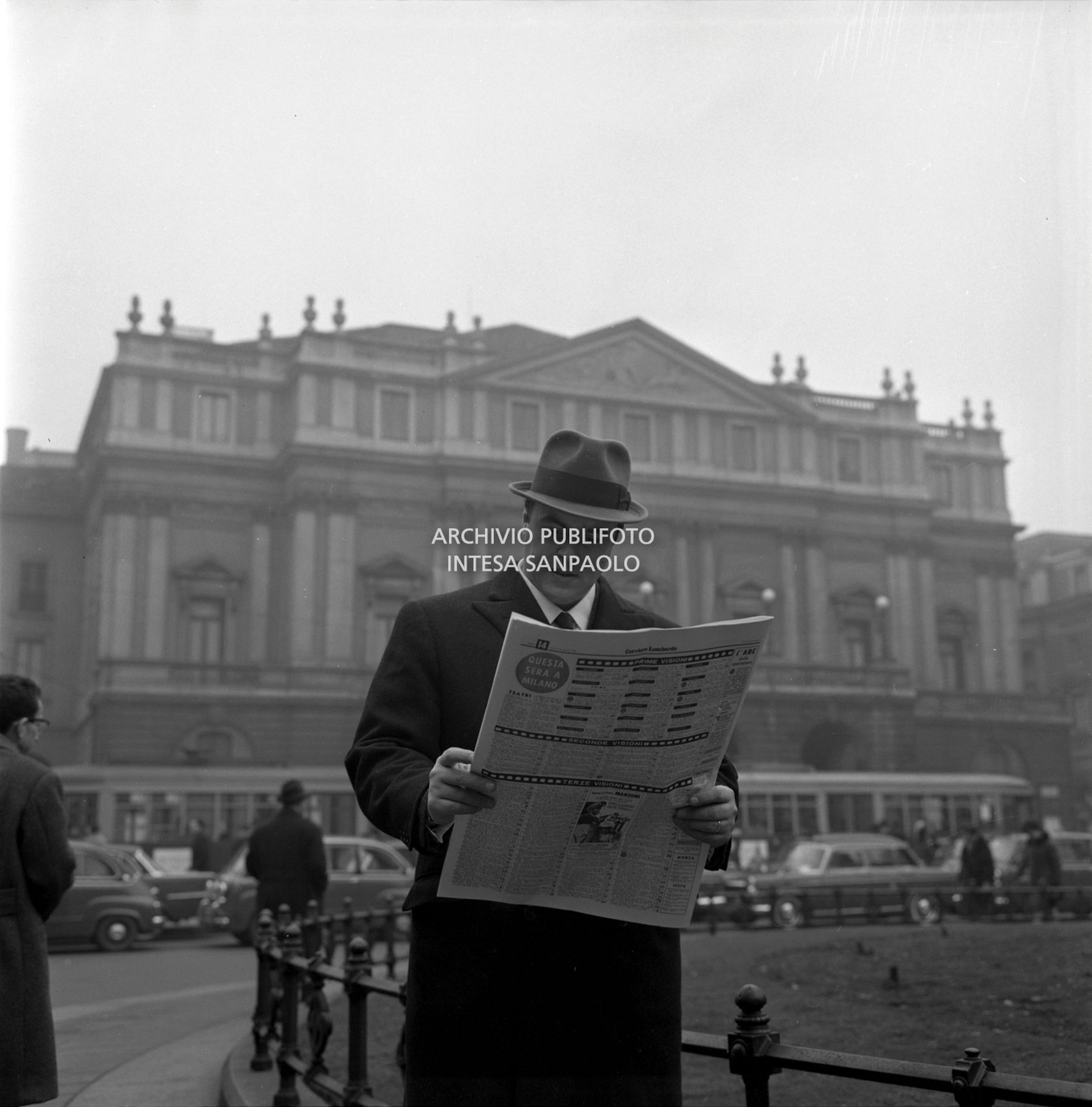 Luciano Pavarotti, ritratto in piazza della Scala a Milano, legge Il Corriere Lombardo; alle sue spalle il Teatro alla Scala
