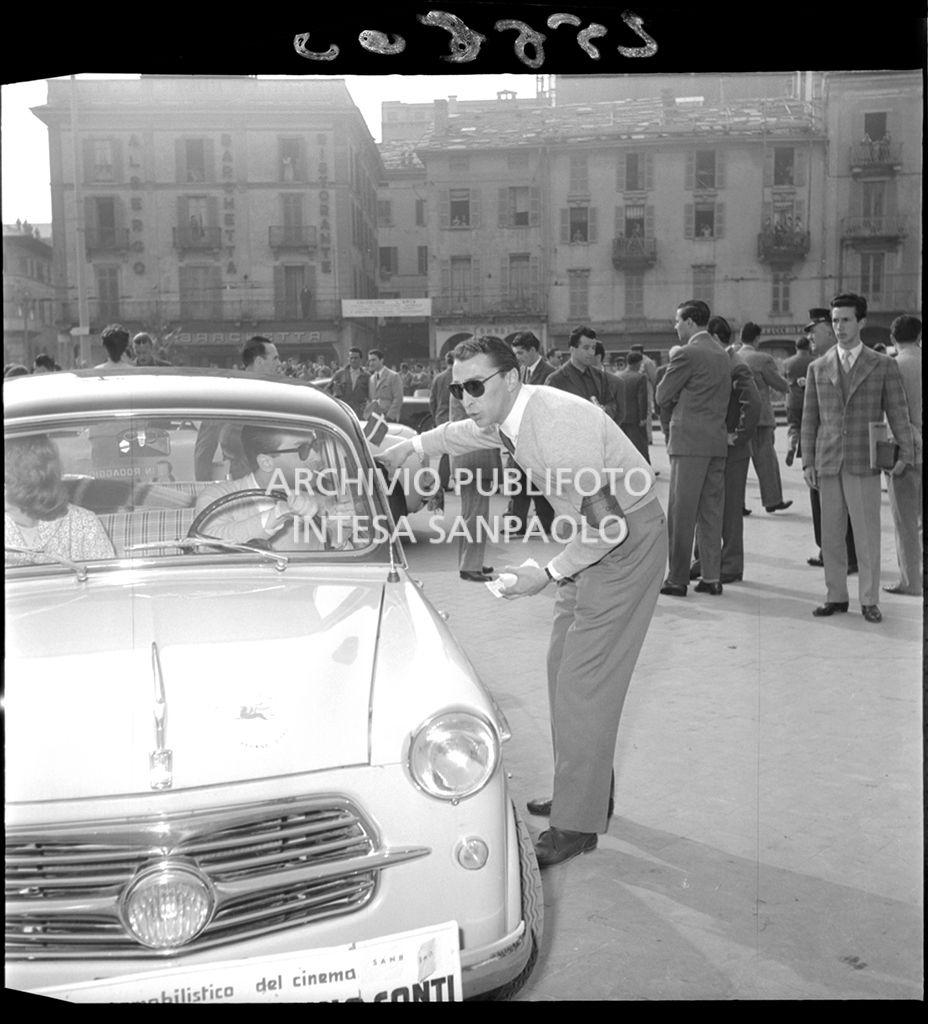 Un membro dell'organizzazione del 2° Rallye automobilistico del cinema in partenza da Como parla con Nino Conti alla guida di una Fiat 1100/103 TV che gareggia insieme a Luciana Angiolillo