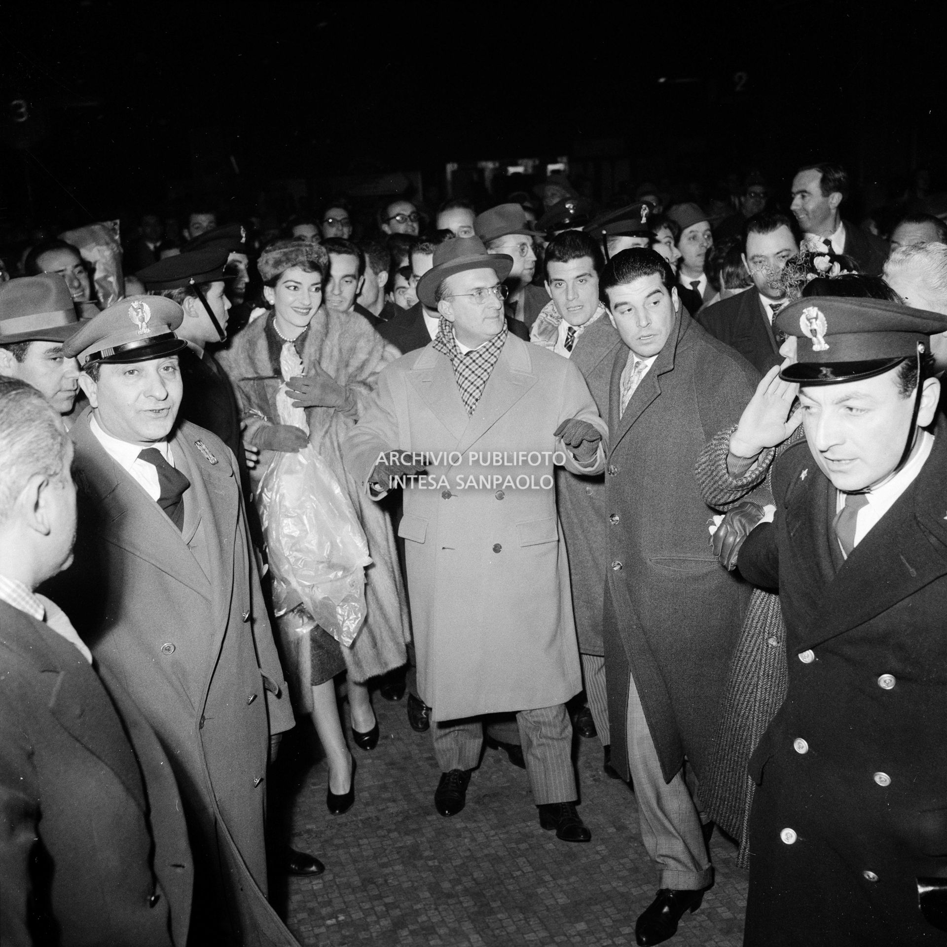 Maria Callas alla Stazione Centrale di Milano, al suo rientro da Roma, si fa strada tra la folla accorsa per accoglierla, con un mazzo di fiori tra le mani