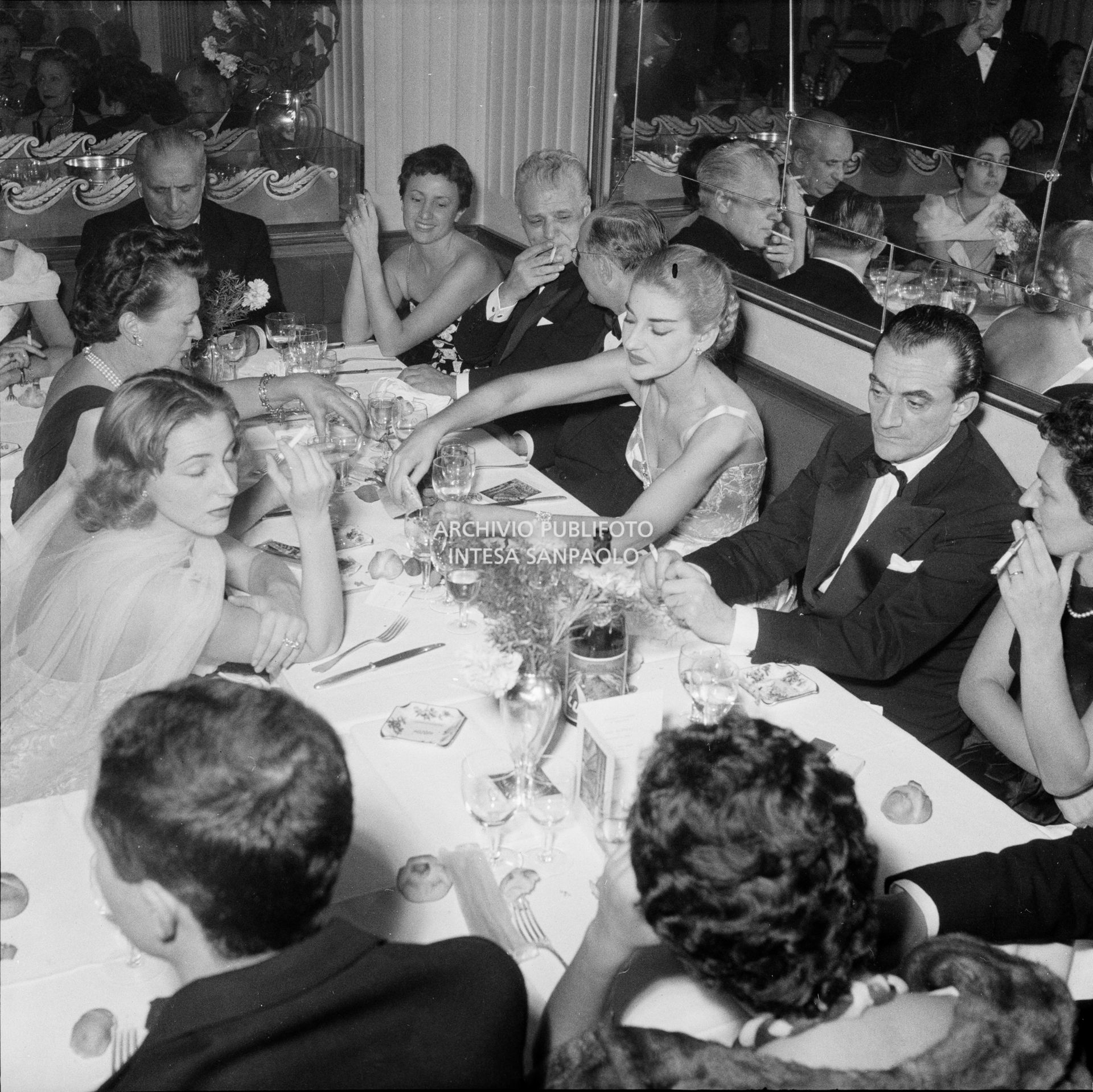 Maria Callas a cena al ristorante Savini di Milano, la sera della Prima della Vestale di Gaspare Spontini. Alla sua sinistra il regista Luchino Visconti, di fronte a lei Emanuela di Castelbarco Toscanini, a capo tavola il marito Giovanni Battista Meneghini, e quinto da destra il soprintendente del Teatro alla Scala Antonio Ghiringhelli
