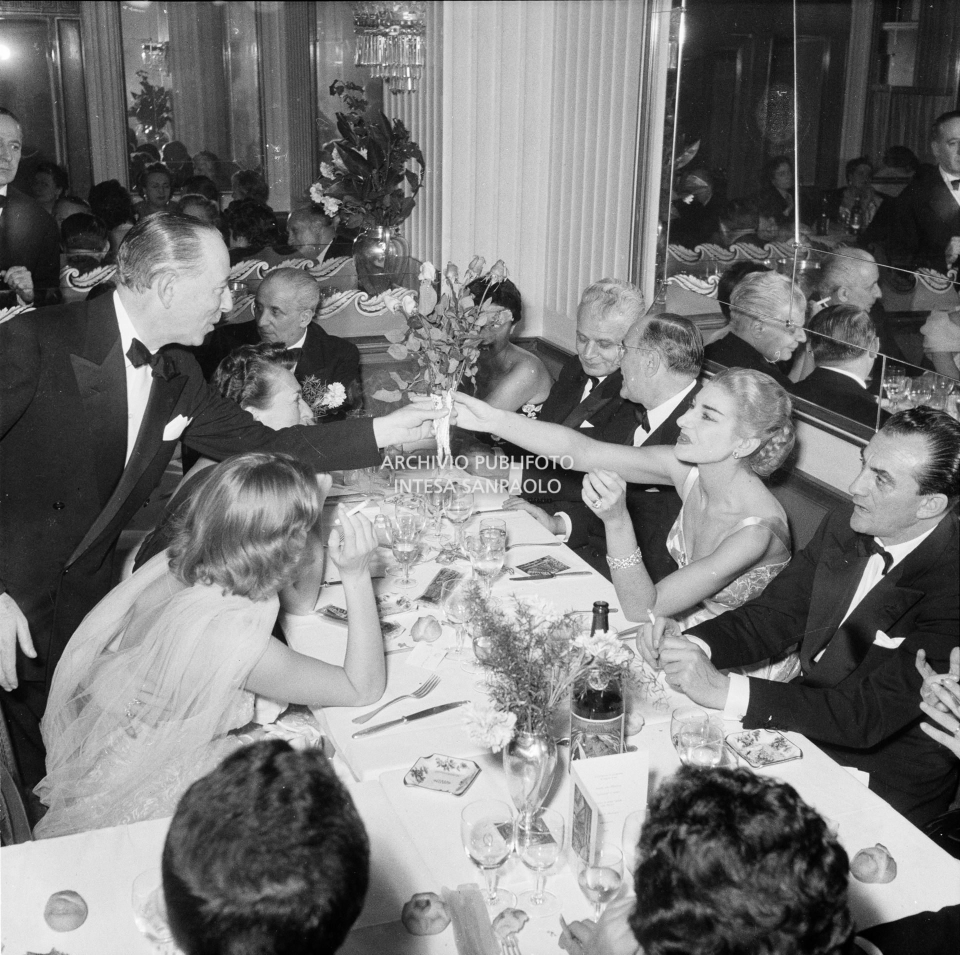 Maria Callas riceve un mazzo di fiori da Valentino Bompiani, al ristorante Savini di Milano, la sera della Prima della Vestale di Gaspare Spontini. Alla sua sinistra il regista Luchino Visconti, a capo tavola il marito Giovanni Battista Meneghini, a sinistra del quale è seduto il soprintendente del Teatro alla Scala Antonio Ghiringhelli