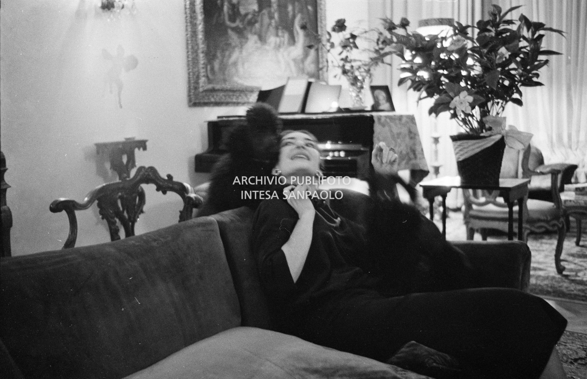 Maria Callas ritratta nella sua abitazione, seduta sul divano in salotto, mentre gioca con il cane