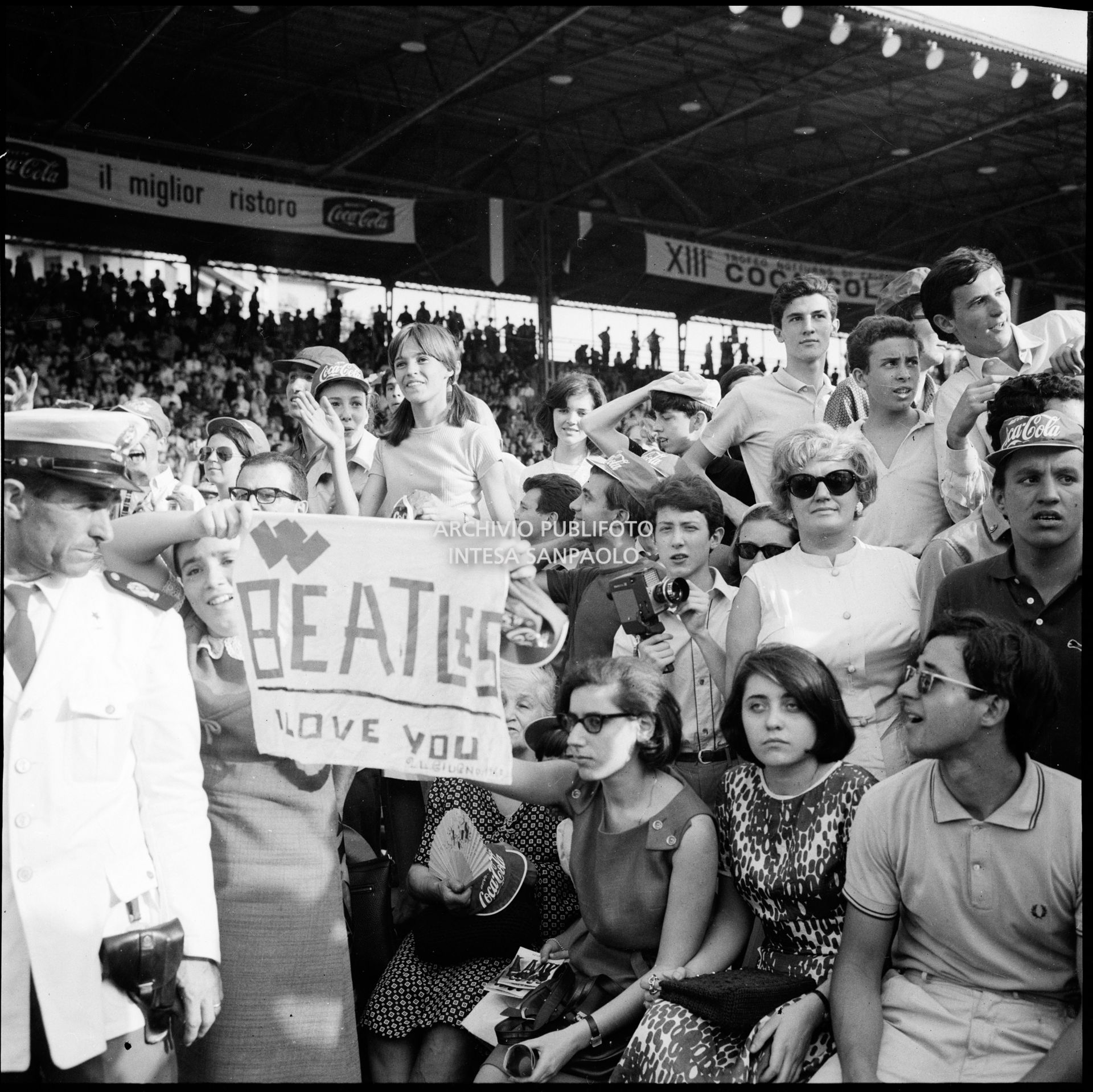Pubblico e forze dell'ordine al concerto pomeridiano dei Beatles al Vigorelli di Milano; una ragazza esibisce un cartello con la scritta: "Beatles I love you".