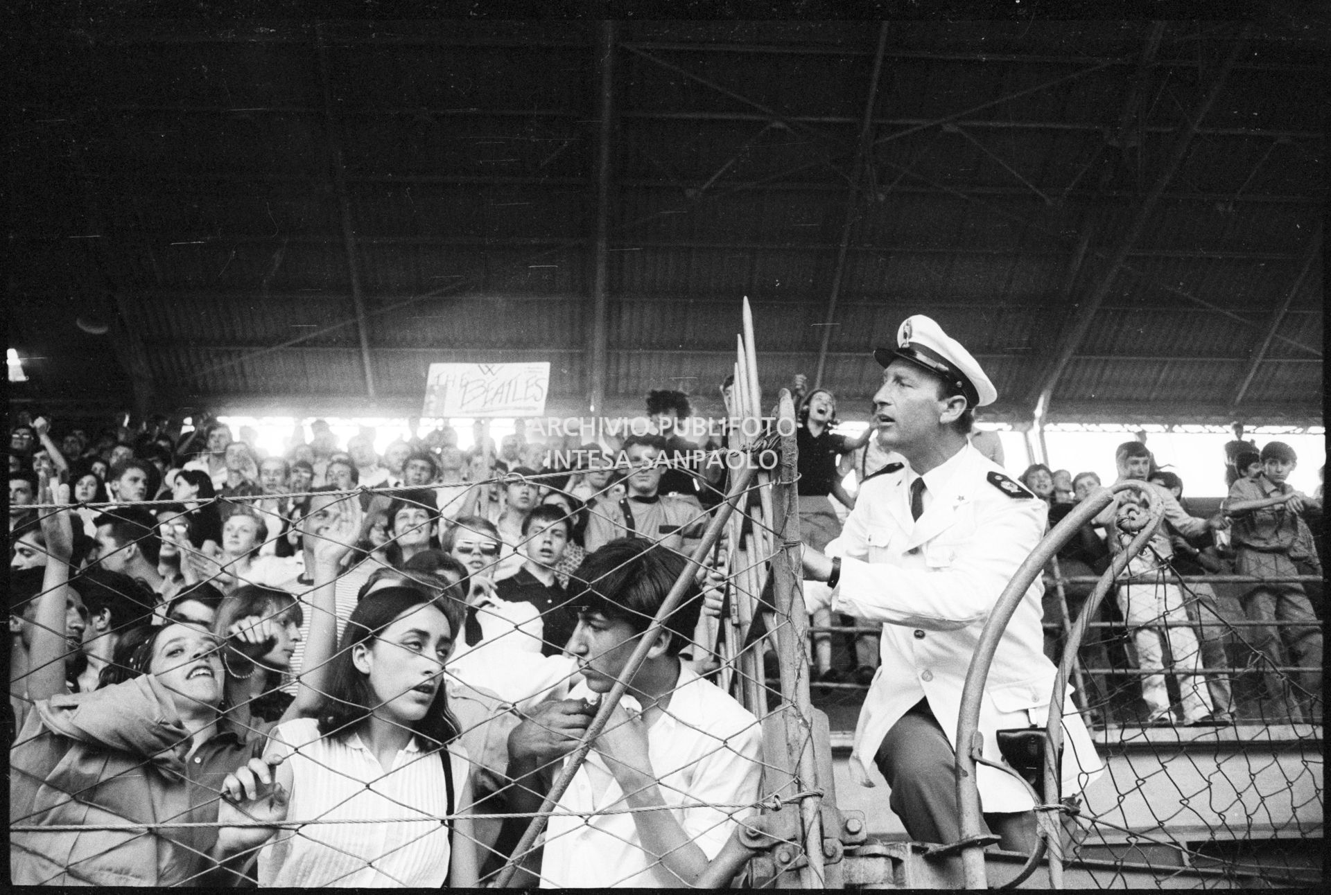 Pubblico sugli spalti al concerto pomeridiano dei Beatles al Vigorelli di Milano; a destra un poliziotto controlla la situazione
