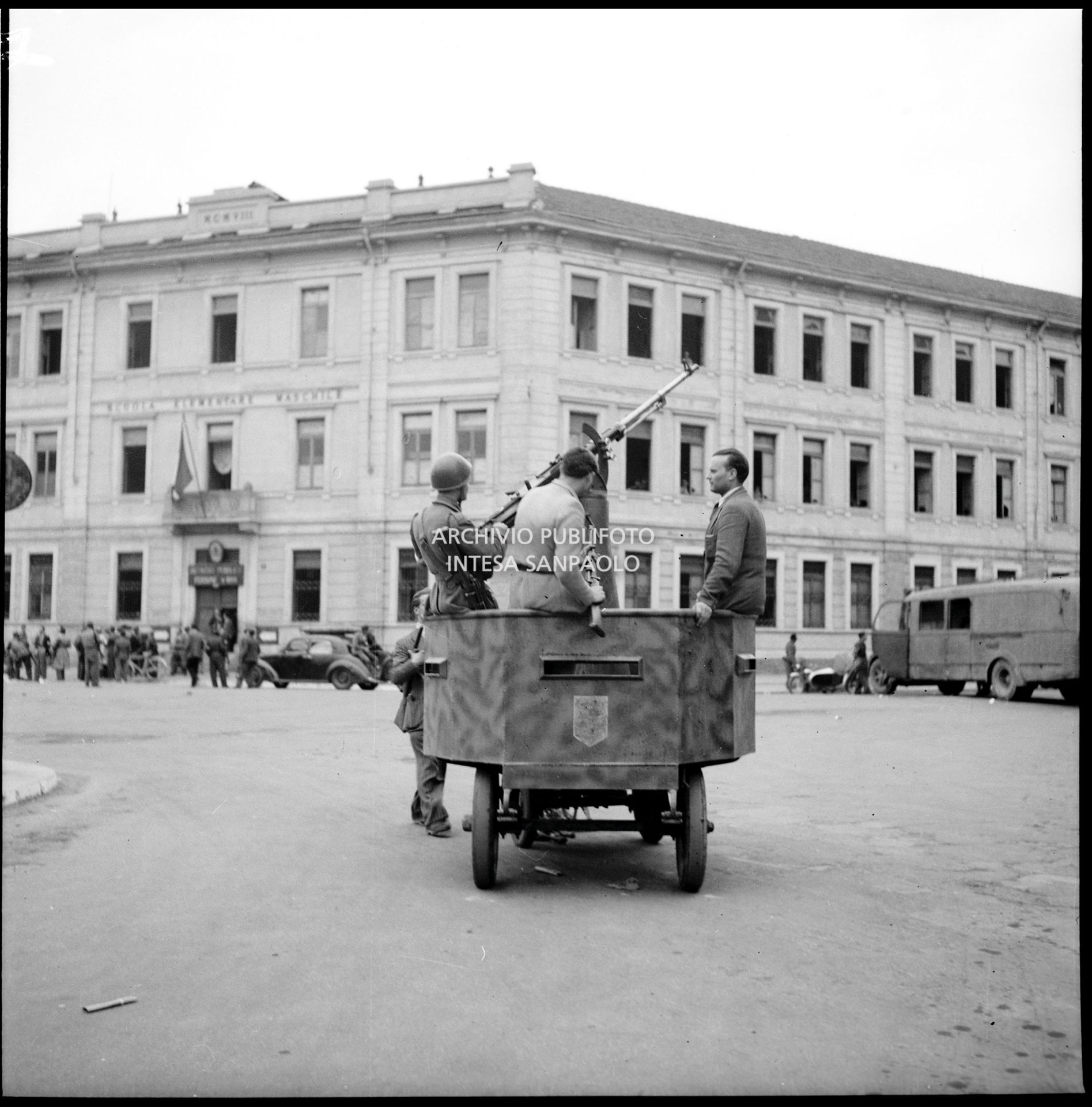 Membri del C.V.L. (Corpo Volontari della Libertà) a bordo di un carrello blindato ed armato con cannoncino contraereo della Legione Autonoma "E. Muti" in piazza Sicilia nella prima giornata dell'insurrezione generale a Milano; sullo sfondo la scuola elementare Nicola Bonservizi convertita in Ricovero pubblico