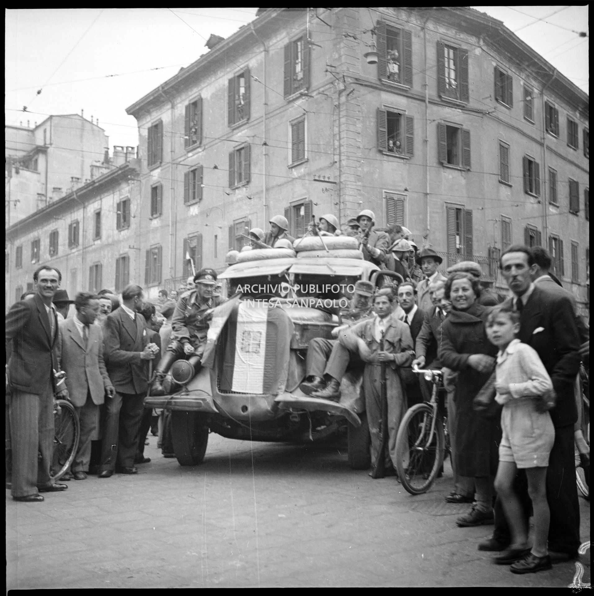Prima giornata di insurrezione generale a Milano: tra la folla, un autocarro con a bordo partigiani, in abiti civili ma con in testa l'elmetto italiano. Sul muso del camion c'è una bandiera italiana con stemma sabaudo posizionata volutamente al contrario; sui parafanghi anteriori sono seduti due soldati tedeschi trasportati come trofeo: a sinistra un generale della Wehrmacht, a destra un soldato
