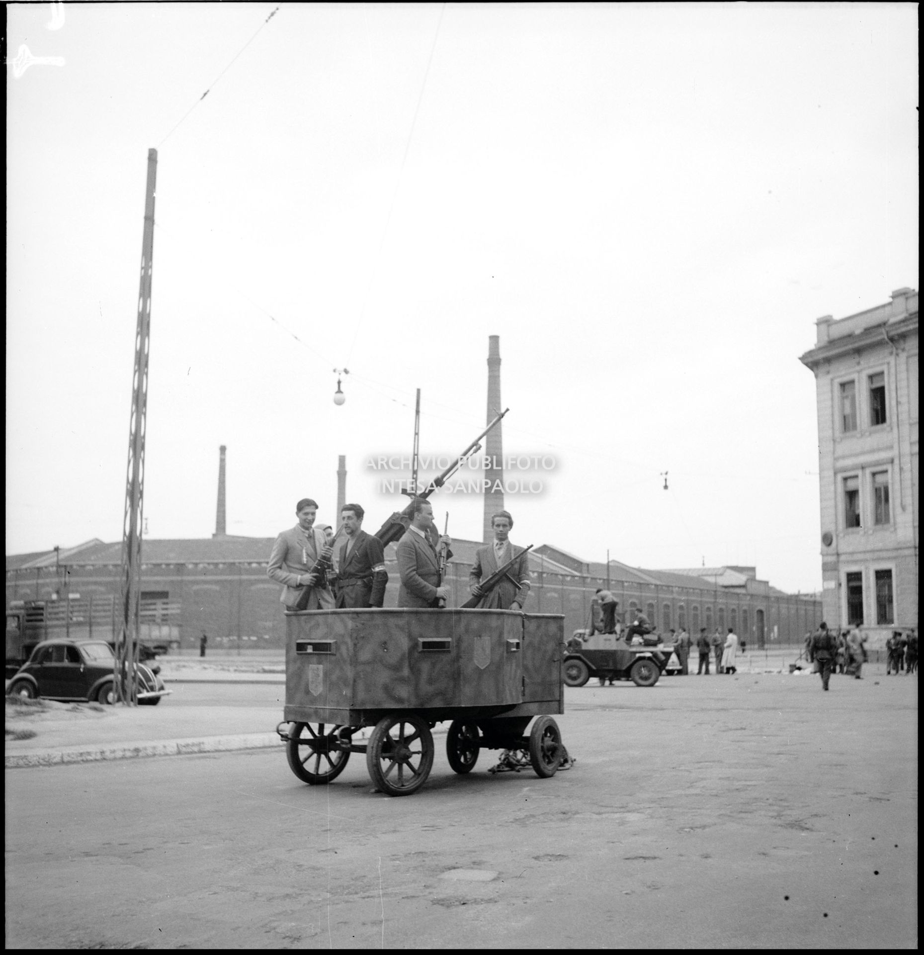 Membri del C.V.L. (Corpo Volontari della Libertà) a bordo di un carrello blindato e armato con cannoncino contraereo della Legione Autonoma "E. Muti" in piazza Sicilia nella prima giornata dell'insurrezione generale a Milano; sullo sfondo la scuola elementare Nicola Bonservizi convertita in Ricovero pubblico