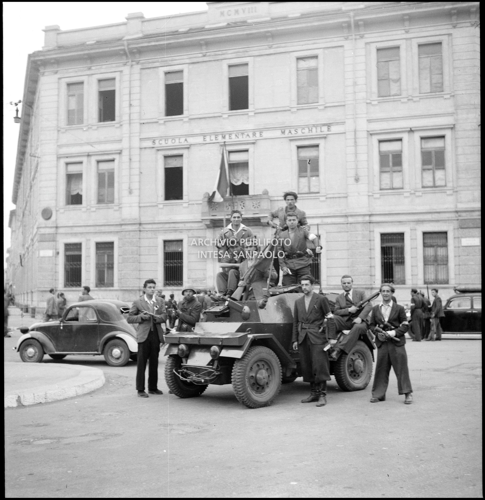 Membri del C.V.L. (Corpo Volontari della Libertà) a bordo di una autoblindo Lancia Ansaldo "Lince" in piazza Sicilia nella prima giornata dell'insurrezione generale a Milano; sullo sfondo la scuola elementare Nicola Bonservizi convertita in Ricovero pubblico
