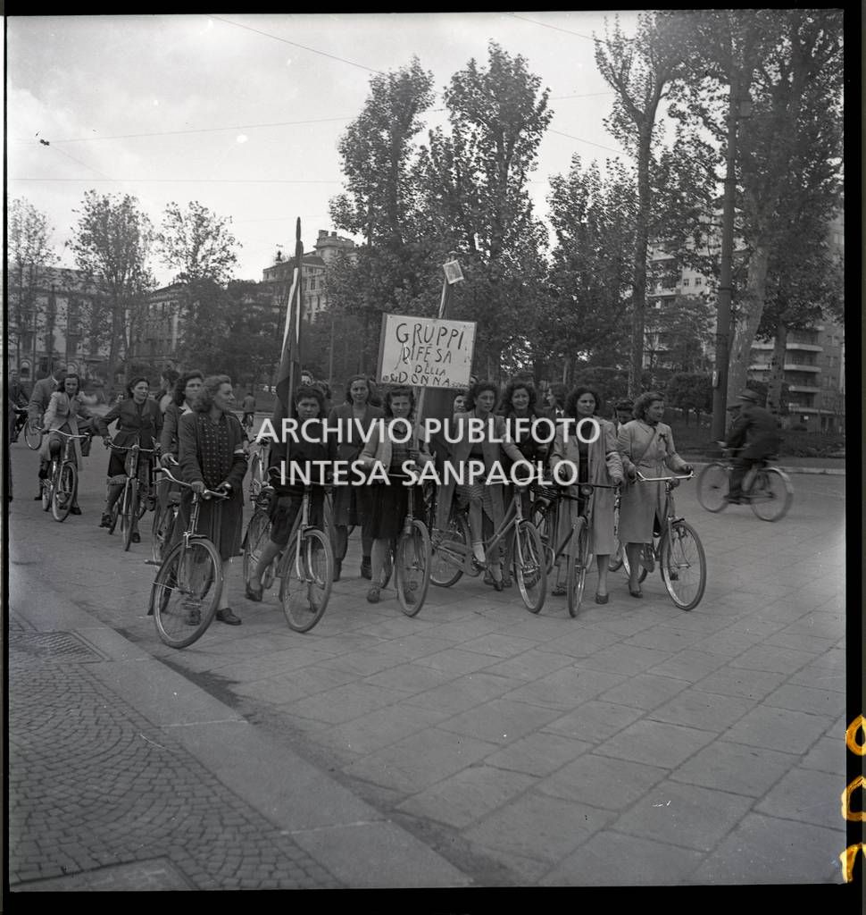 Donne dei Gruppi di difesa della donna (GDD) in bicicletta con bandiere e un cartello che recita: Gruppi di Difesa della Donna