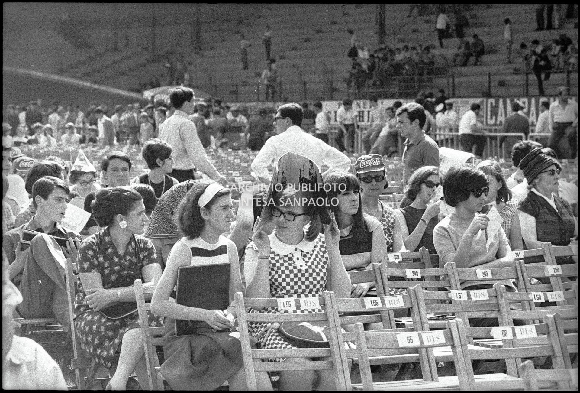 Pubblico sotto il sole al Vigorelli di Milano in attesa dell'inizio del concerto pomeridiano dei Beatles; una ragazza si ripara con il pieghevole del programma