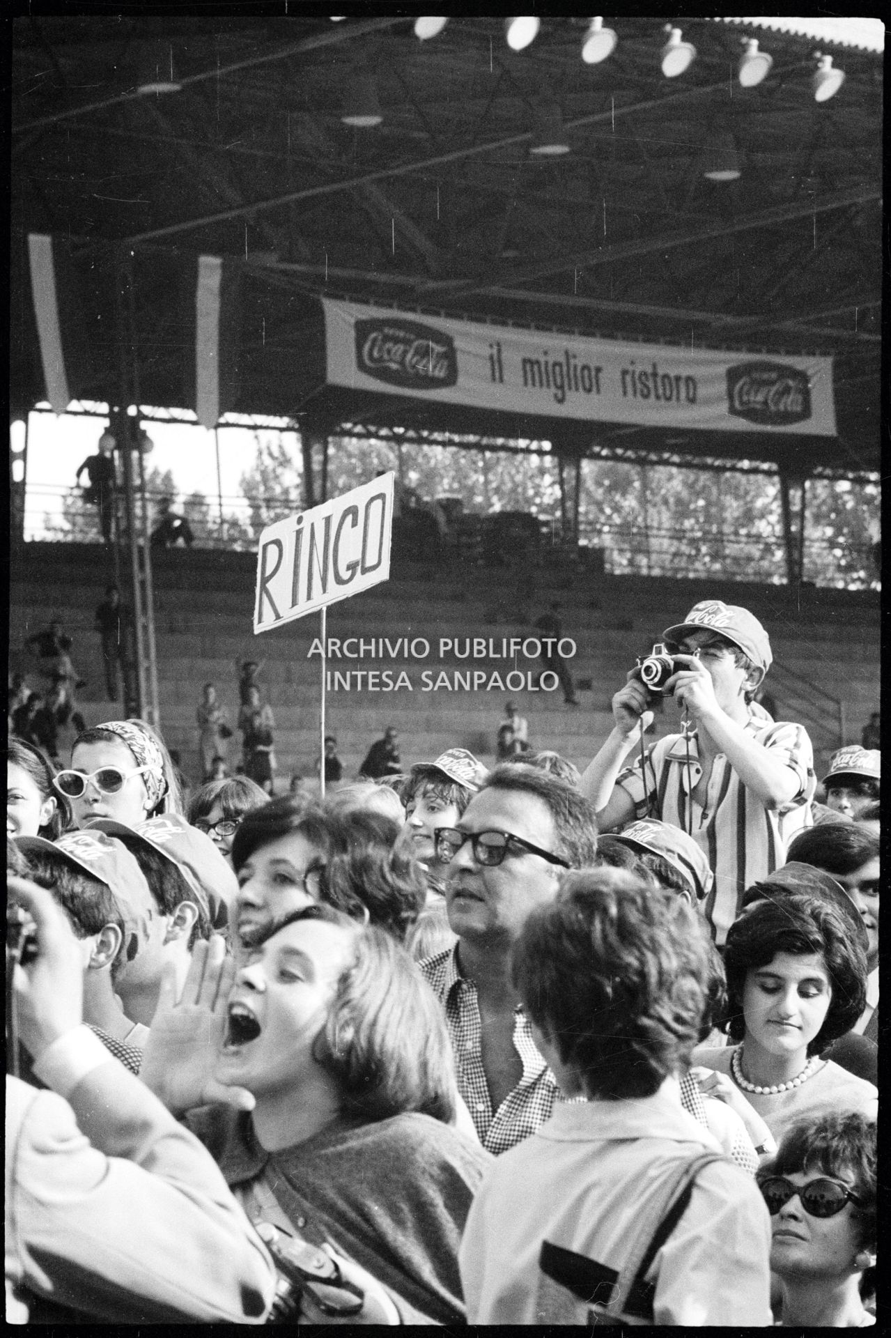 Pubblico al concerto pomeridiano dei Beatles al Vigorelli di Milano; un ragazzo con la macchina fotografica, un'altra tiene un cartello con la scritta "Ringo"