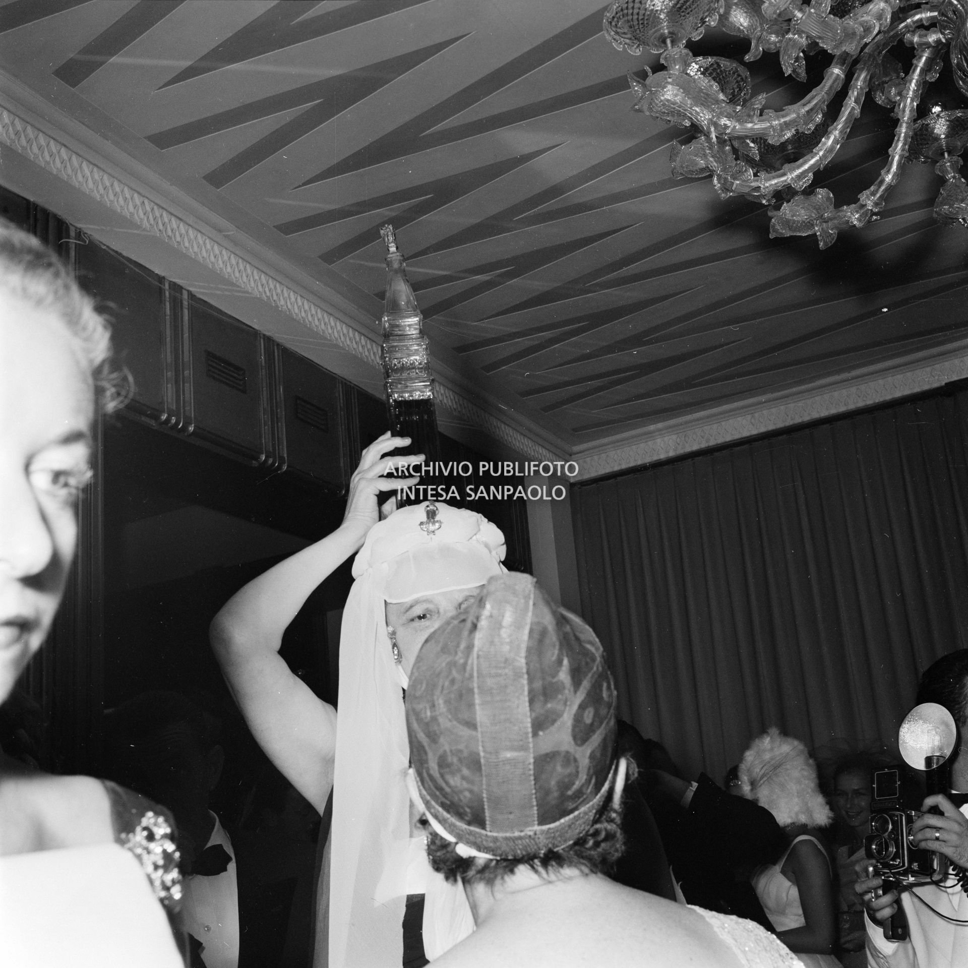 La marchesa Olga Cadaval, con la bottiglia a forma di campanile di San Marco in testa, con Elsa Maxwell (di spalle), tra gli altri, al ricevimento organizzato in onore di Maria Callas