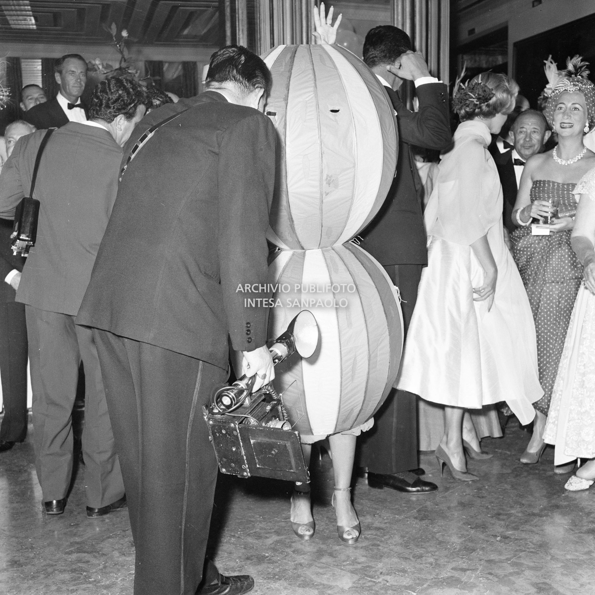 Un fotografo sbircia nel costume che avvolge Miss Maria Salata Philips al ricevimento organizzato da Elsa Maxwell in onore di Maria Callas