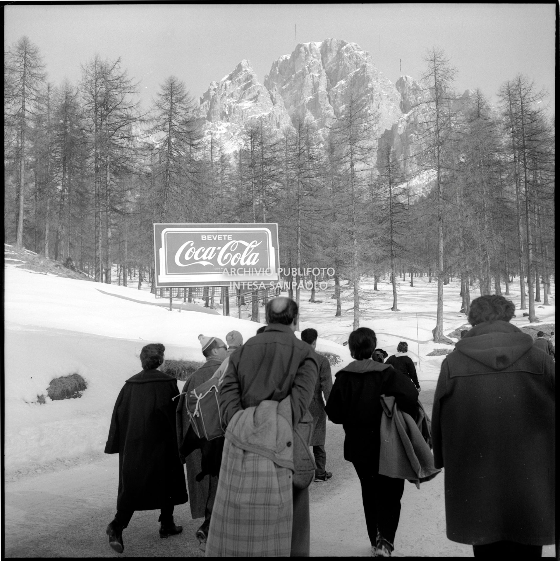 Cartello pubblicitario Coca-Cola lungo una strada, affollata di passanti, nei pressi di Cortina d'Ampezzo nel periodo dei VII Giochi olimpici invernali