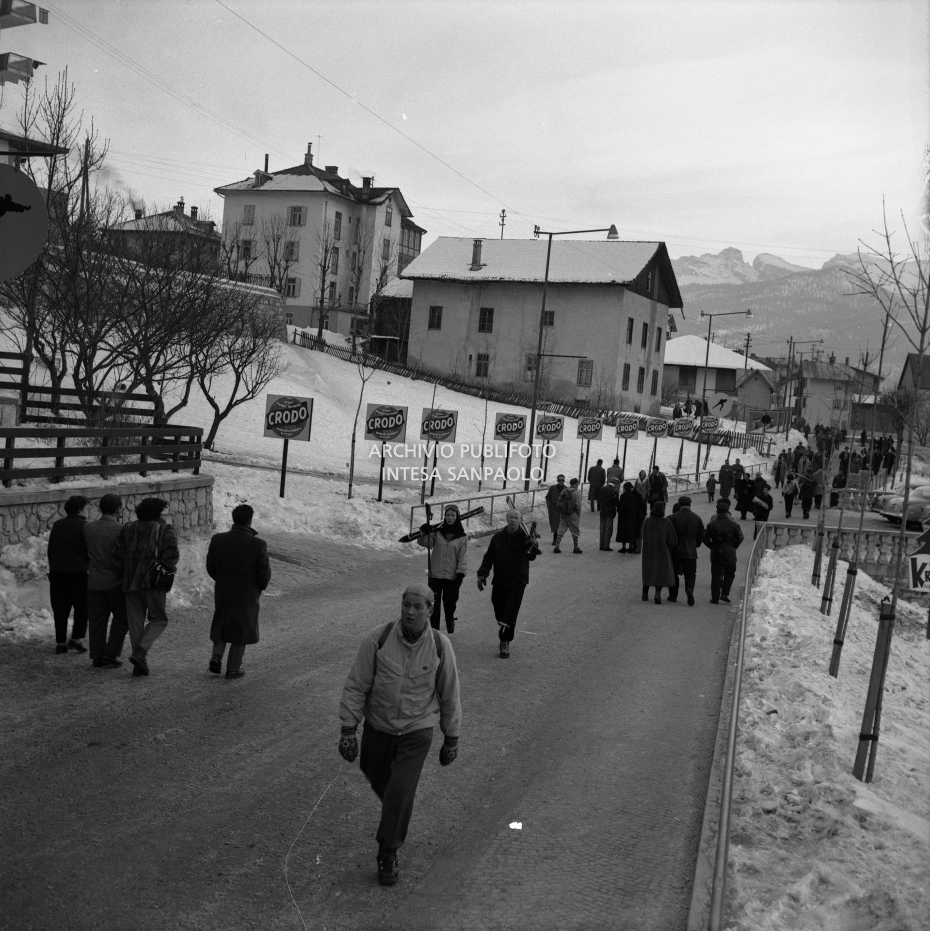 Cartelli pubblicitari dell'acqua e delle bibite Crodo lungo via del Castello, percorsa da alcuni passanti, a Cortina d'Ampezzo nel periodo dei VII Giochi olimpici invernali