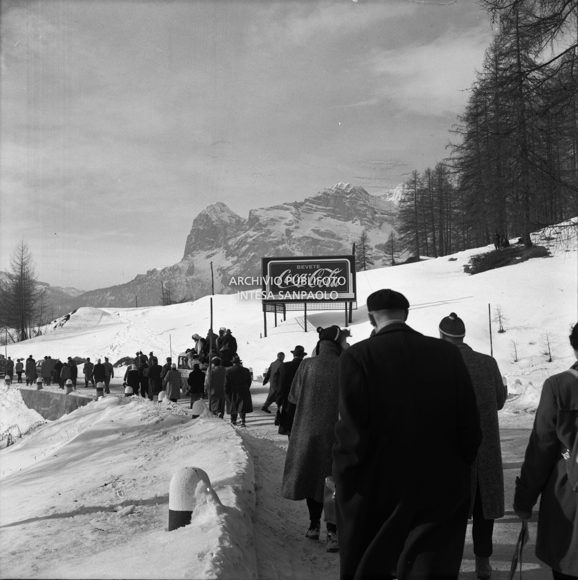 Cartello pubblicitario Coca-Cola lungo una strada, affollata di passanti, nei pressi di Cortina d'Ampezzo nel periodo dei VII Giochi olimpici invernali