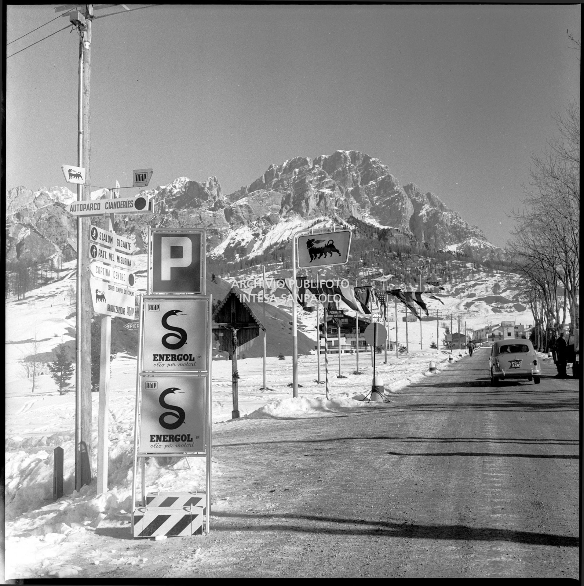 Cartelli dell'olio motore Agip Energol sulla strada regionale delle Dolomiti imbandierata nei pressi di Cortina d'Ampezzo nel periodo dei VII Giochi olimpici invernali