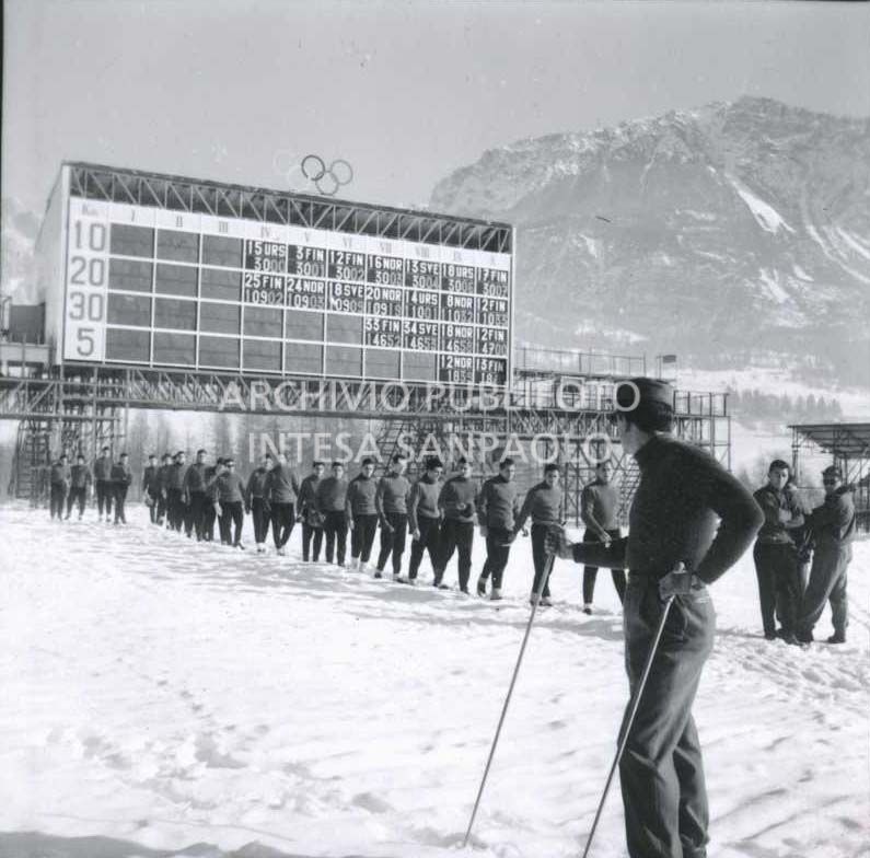 Il tabellone dei tempi delle gare di fondo presso lo stadio della neve; in primo piano alcuni atleti italiani