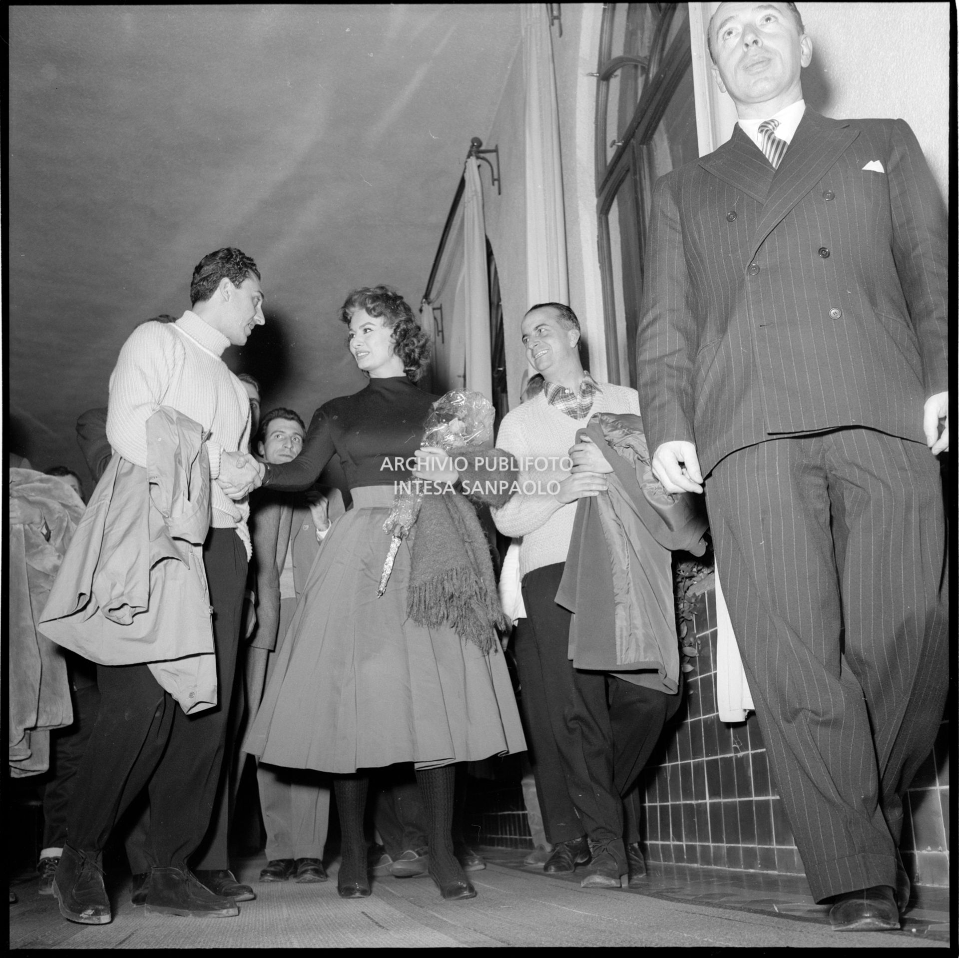 Sophia Loren al suo arrivo a Cortina d'Ampezzo in occasione dei VII Giochi olimpici invernali con un mazzo di fiori in mano stringe la mano a un uomo