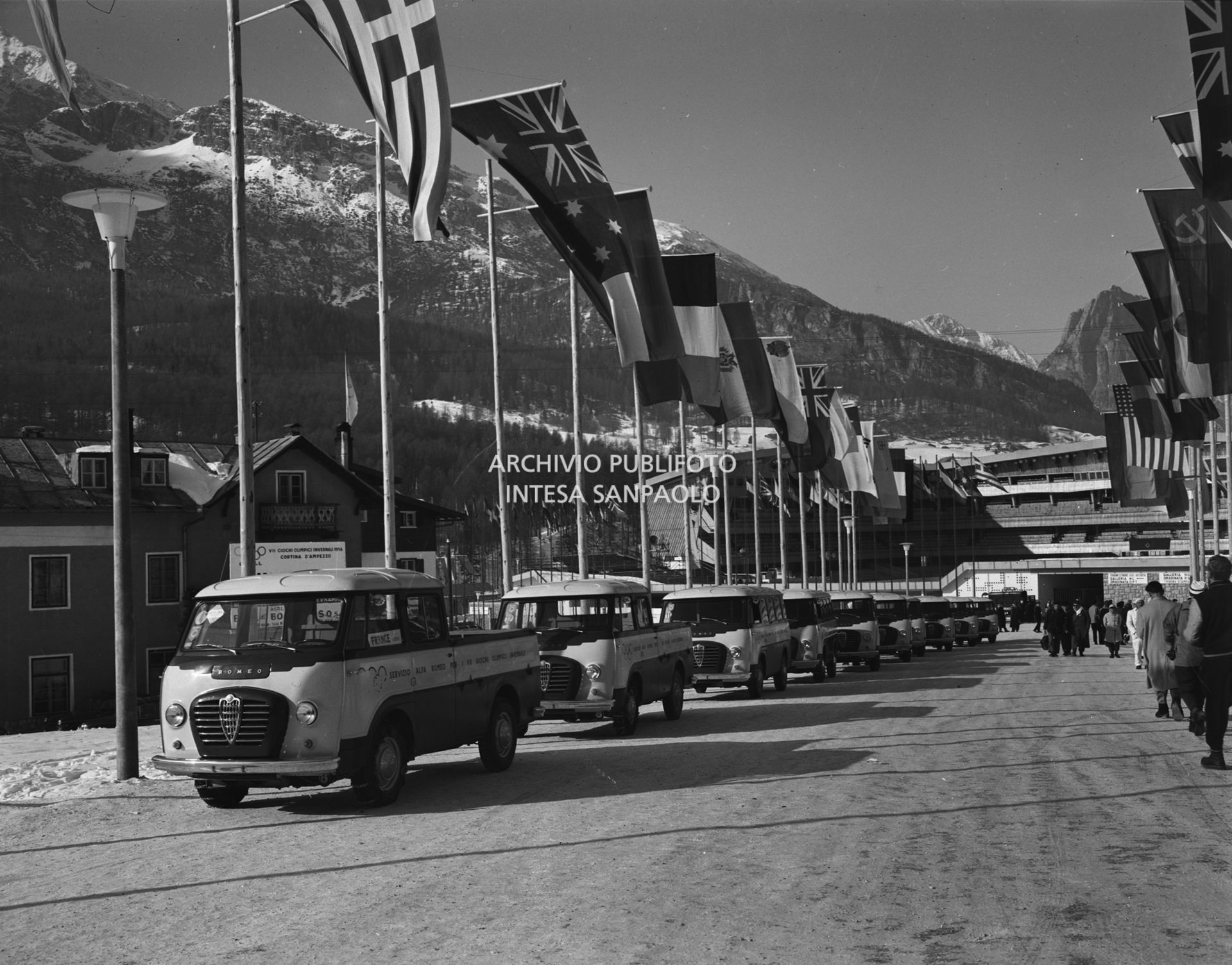 Furgoni Romeo della Alfa Romeo, a disposizione dei partecipanti ai VII Giochi olimpici invernali, in colonna nel viale di ingresso allo Stadio del ghiaccio, a Cortina d'Ampezzo, decorato con le bandiere dei vari Paesi