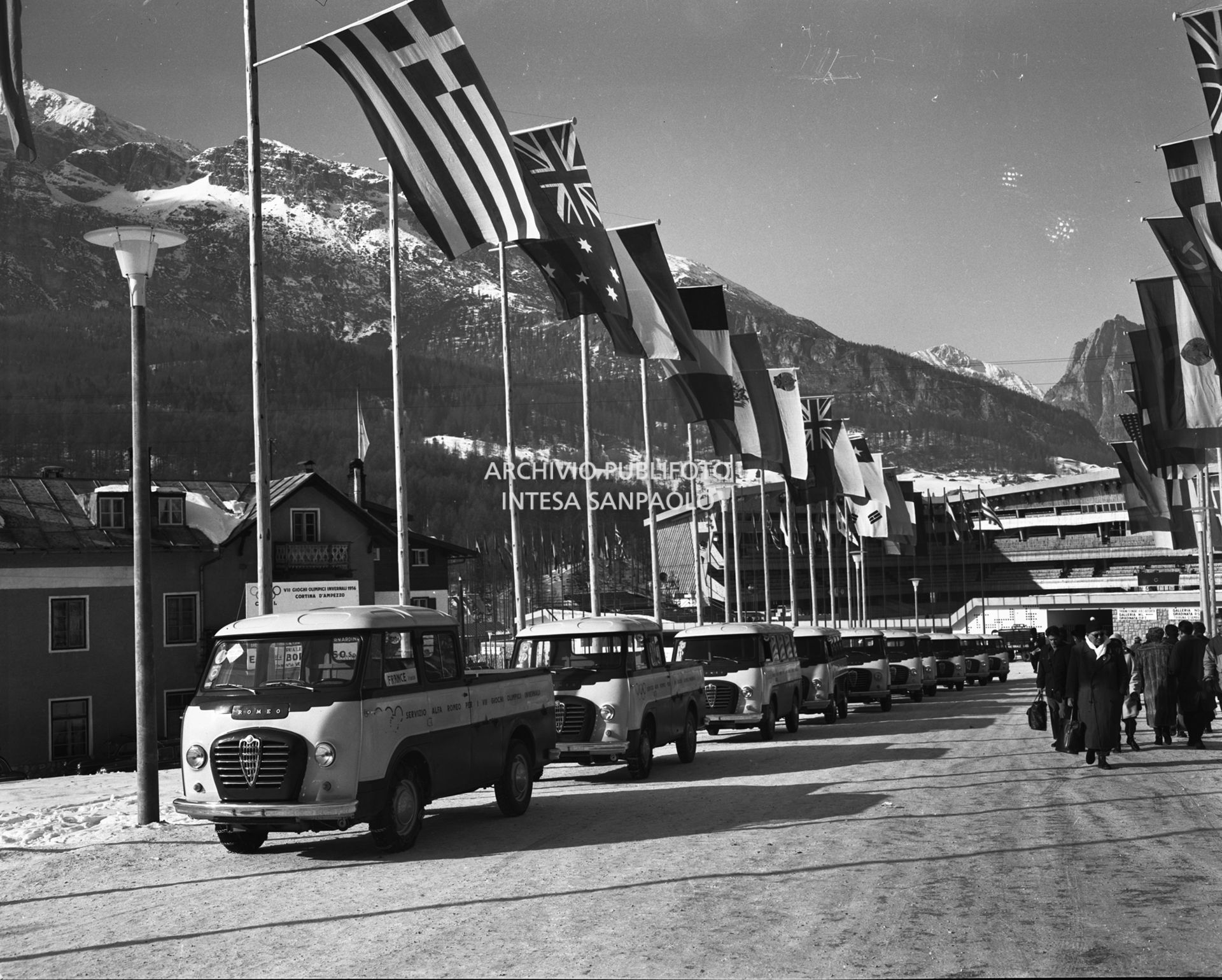 Furgoni Romeo della Alfa Romeo, a disposizione dei partecipanti ai VII Giochi olimpici invernali, in colonna nel viale di ingresso allo Stadio del ghiaccio, a Cortina d'Ampezzo, decorato con le bandiere dei vari Paesi