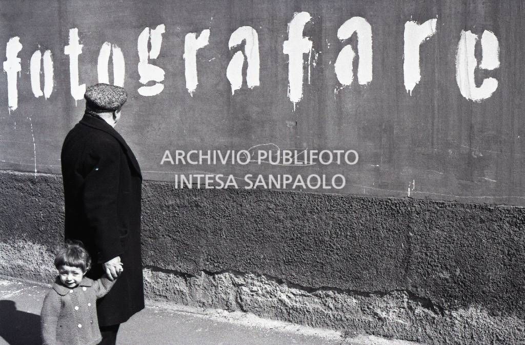 Un uomo con una bambina per mano osserva la scritta "Fotografare" dipinta su un muro