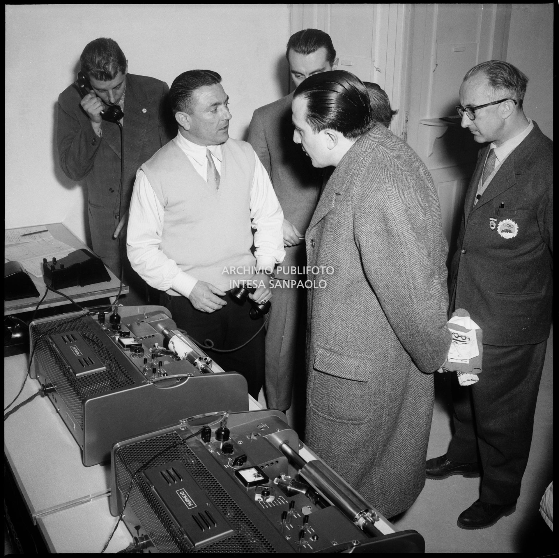 Il ministro delle Finanze Giulio Andreotti visita il reparto delle telefoto del Centro per la stampa ospitato all'albergo Savoia di Cortina d'Ampezzo; alle sue spalle il direttore del Centro Giuseppe Sabelli Fioretti