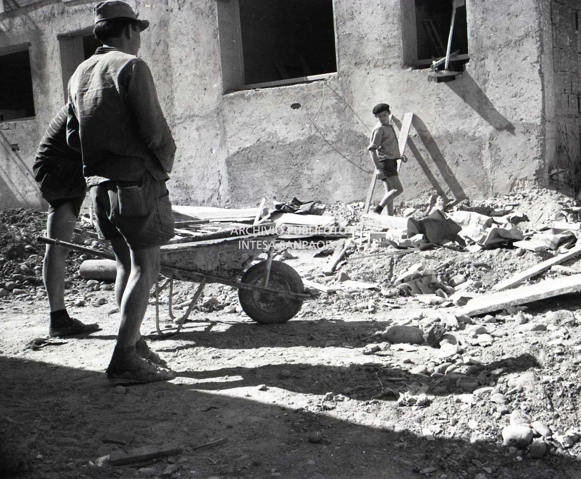 Ragazzino al lavoro in un cantiere edile: in primo piano due altri manovali con carriola
