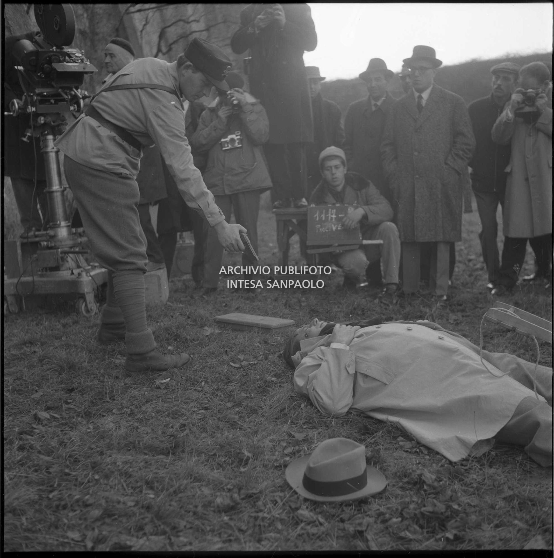 Una scena del film "Il processo di Verona" in cui viene interpretata una fucilazione; alle spalle degli attori è visibile un uomo con un ciak