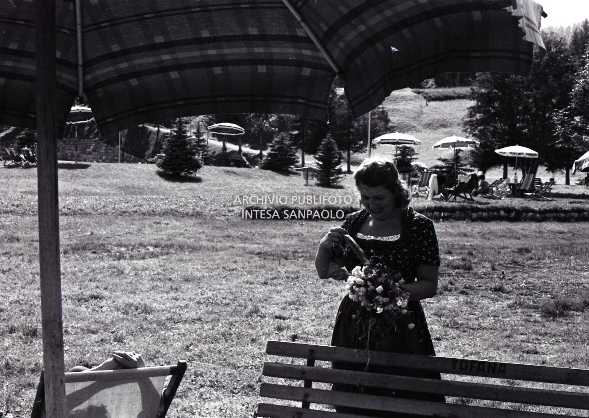 Donna con un mazzo di fiori in un prato a Cortina d'Ampezzo ; sullo sfondo ombrelloni e sdraio