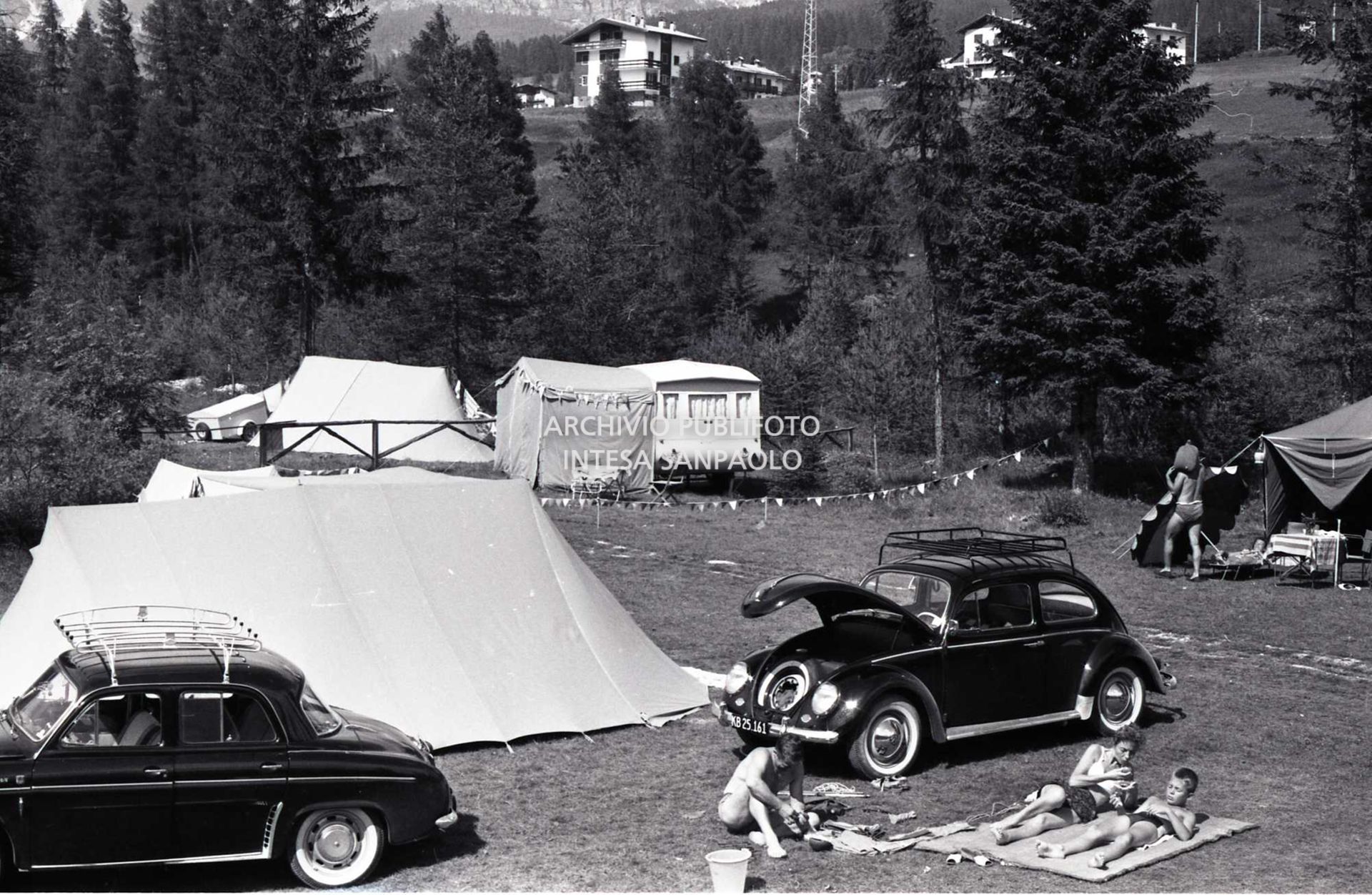 Turisti in campeggio a Cortina d'Ampezzo sdraiati in un prato a fianco a un Maggiolino Volkswagen ; sulla sinistra una Renault Dauphine/Ondine