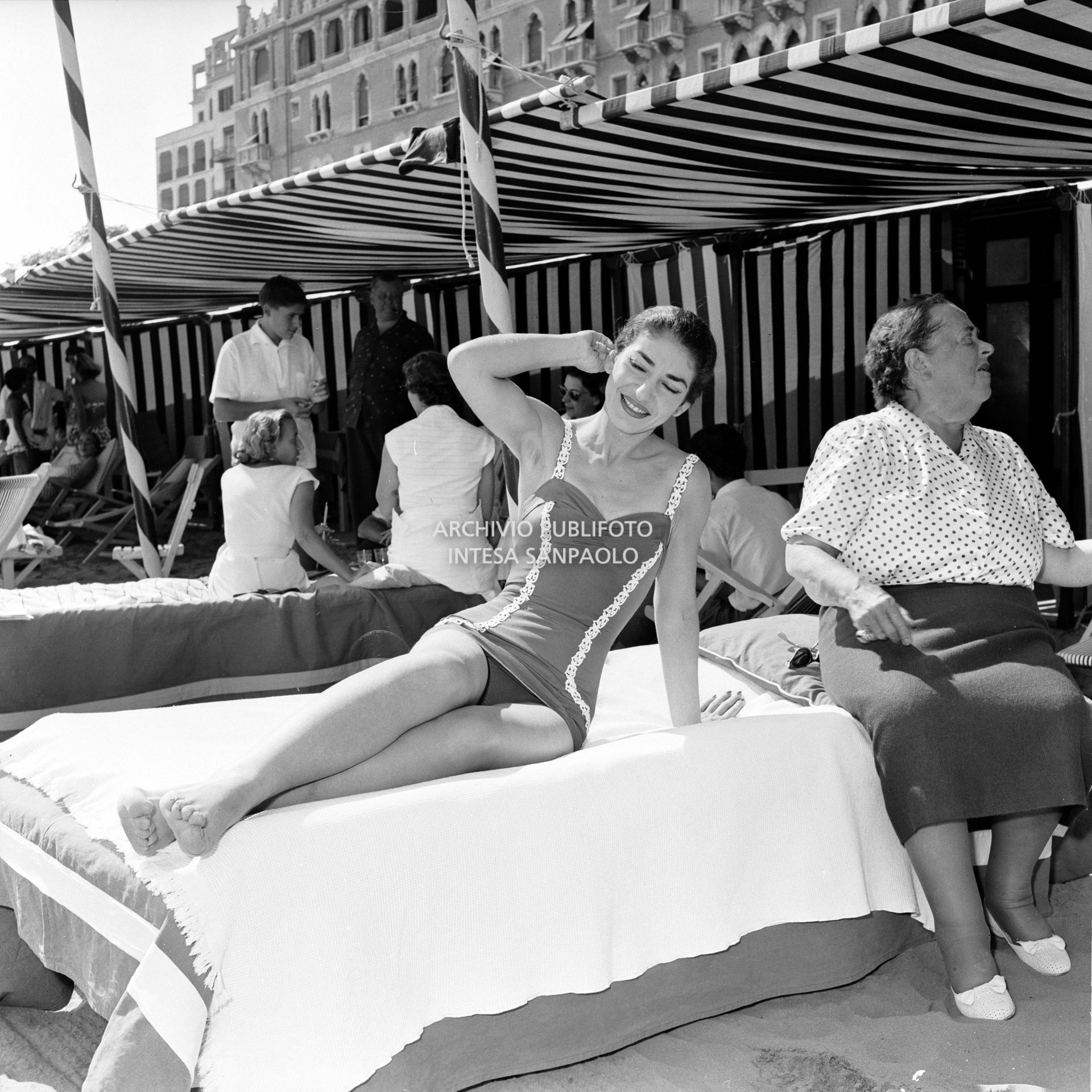 La soprano Maria Callas in costume da bagno sdraiata su un lettino al Lido di Venezia; a destra Elsa Maxwell