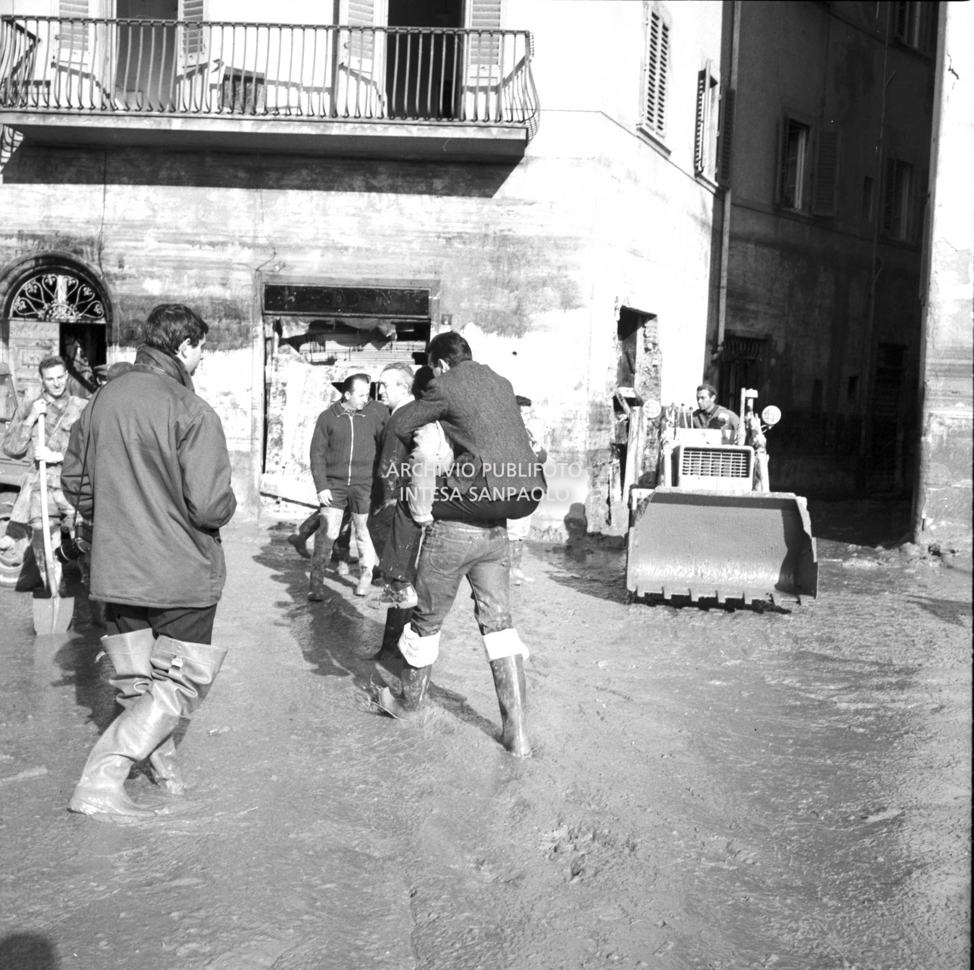 Alluvione di Firenze: piazza Mentana invasa dal fango, un uomo viene trasportato sulla schiena, sullo sfondo una ruspa