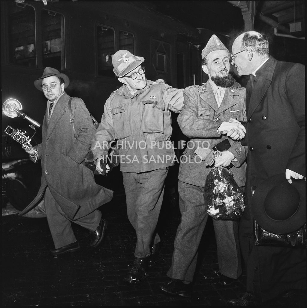 Padre Brevi di rientro dalla prigionia in Unione Sovietica cammina sulla banchina di una stazione ferroviaria, accolto da un sacerdote e un militare. Sulla sinistra un fotografo<br>196452
