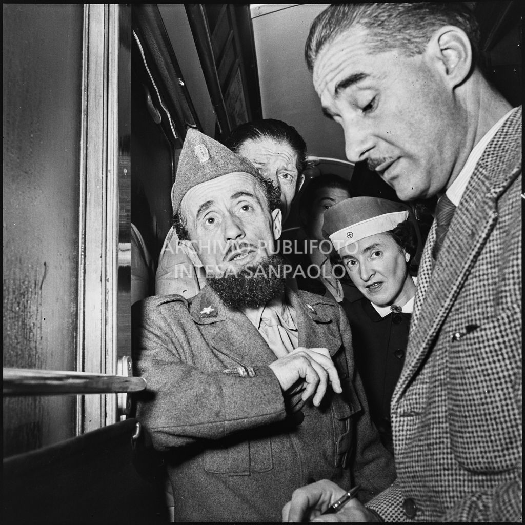 Padre Brevi di rientro in patria dopo la prigionia in Unione Sovietica intervistato da un giornalista a bordo di un treno. Alle sue spalle altri uomini e un'infermiera<br>196458