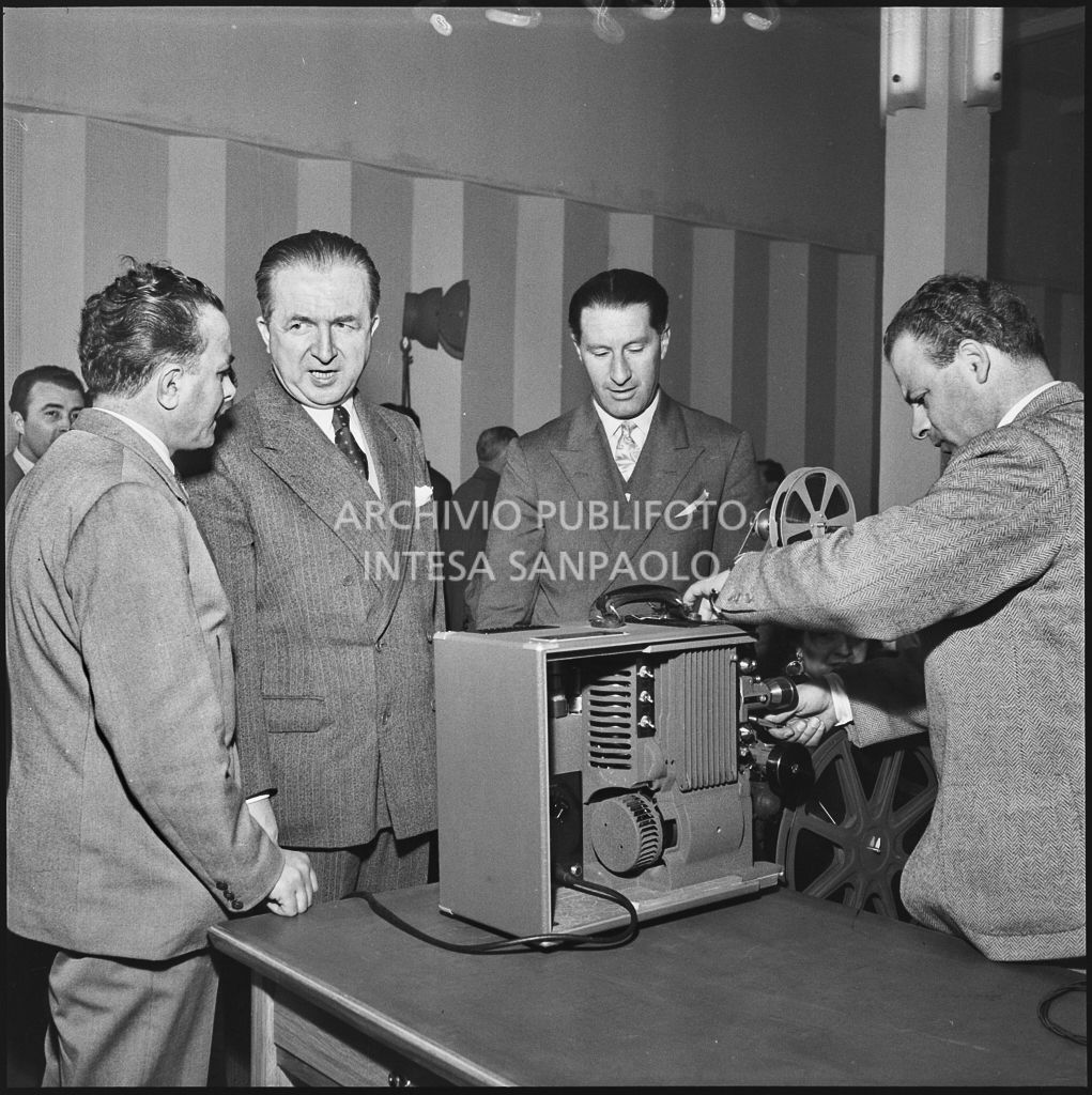 L'onorevole Giuseppe Pella osserva un uomo mentre prepara un proiettore in occasione del convegno "Human relations" nella sala Puricelli della Fiera campionaria di Milano del 1954<br>210823