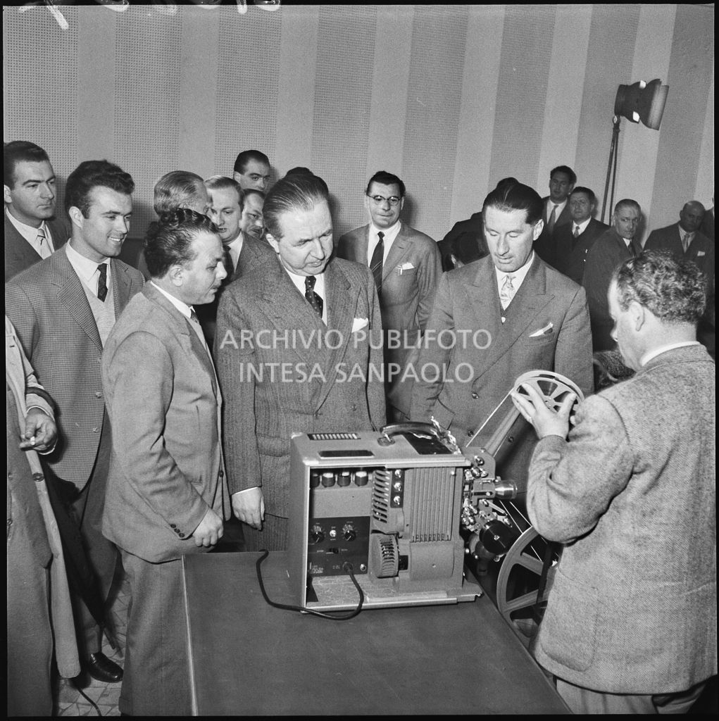 L'onorevole Giuseppe Pella osserva un uomo mentre prepara un proiettore in occasione del convegno "Human relations" nella sala Puricelli della Fiera campionaria di Milano del 1954<br>210824