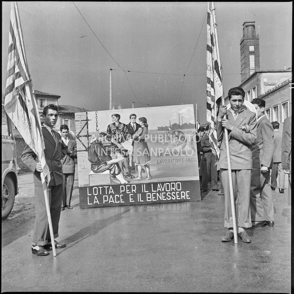 Due ragazzi sorreggono delle bandiere davanti ad un pannello che recita "Lotta per il lavoro la pace e il benessere" in occasione della Festa del 1° maggio<br>212215