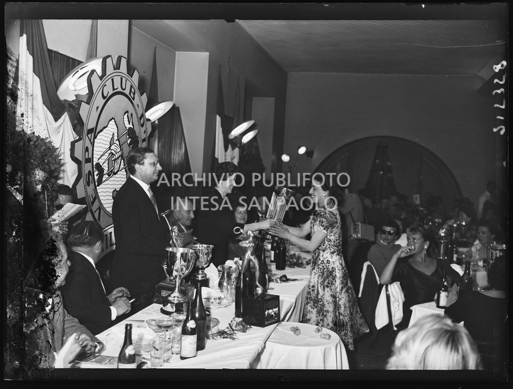 Una donna consegna un premio ad una delle vincitrici della terza edizione dell'Audax internazionale vespistico femminile durante il banchetto organizzato del Vespa Club Milano. In piedi dietro al tavolo il presidente del Vespa Club Milano Renato Tassinari<br>215358