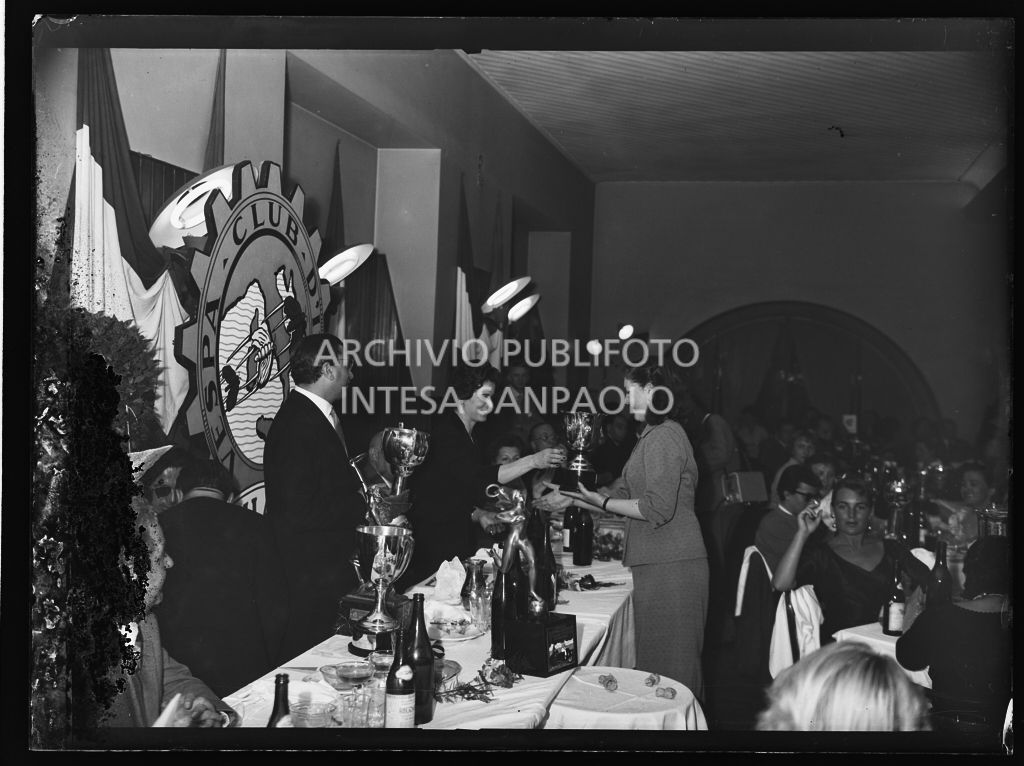 Una donna consegna un premio ad una delle vincitrici della terza edizione dell'Audax internazionale vespistico femminile durante il banchetto organizzato del Vespa Club Milano. In piedi dietro al tavolo il presidente del Vespa Club Milano Renato Tassinari<br>215359