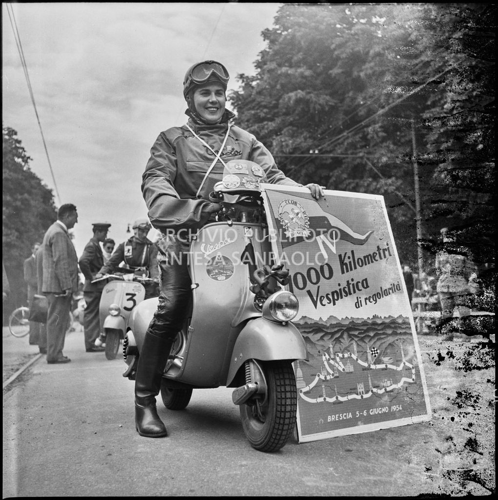 La concorrente Ada Pace del Vespa Club di Torino (numero di gara 1) in sella alla sua Vespa e con in mano il cartellone della competizione sportiva 1000 Km Vespistica del 1954<br>216206