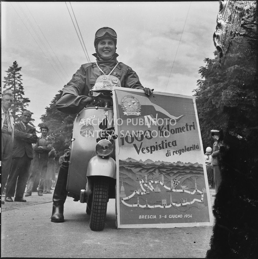 La concorrente Ada Pace del Vespa Club di Torino (numero di gara 1) in sella alla sua Vespa e con in mano il cartellone della competizione sportiva 1000 Km Vespistica del 1954<br>216306