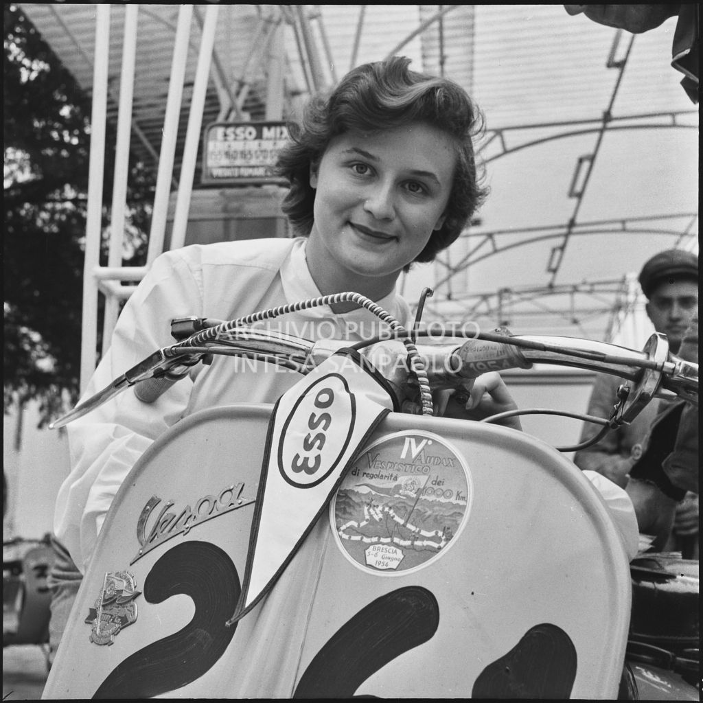 Una donna fotografata accanto ad una Vespa della competizione sportiva 1000 Km Vespistica del 1954 durante una pausa rifornimento<br>216383