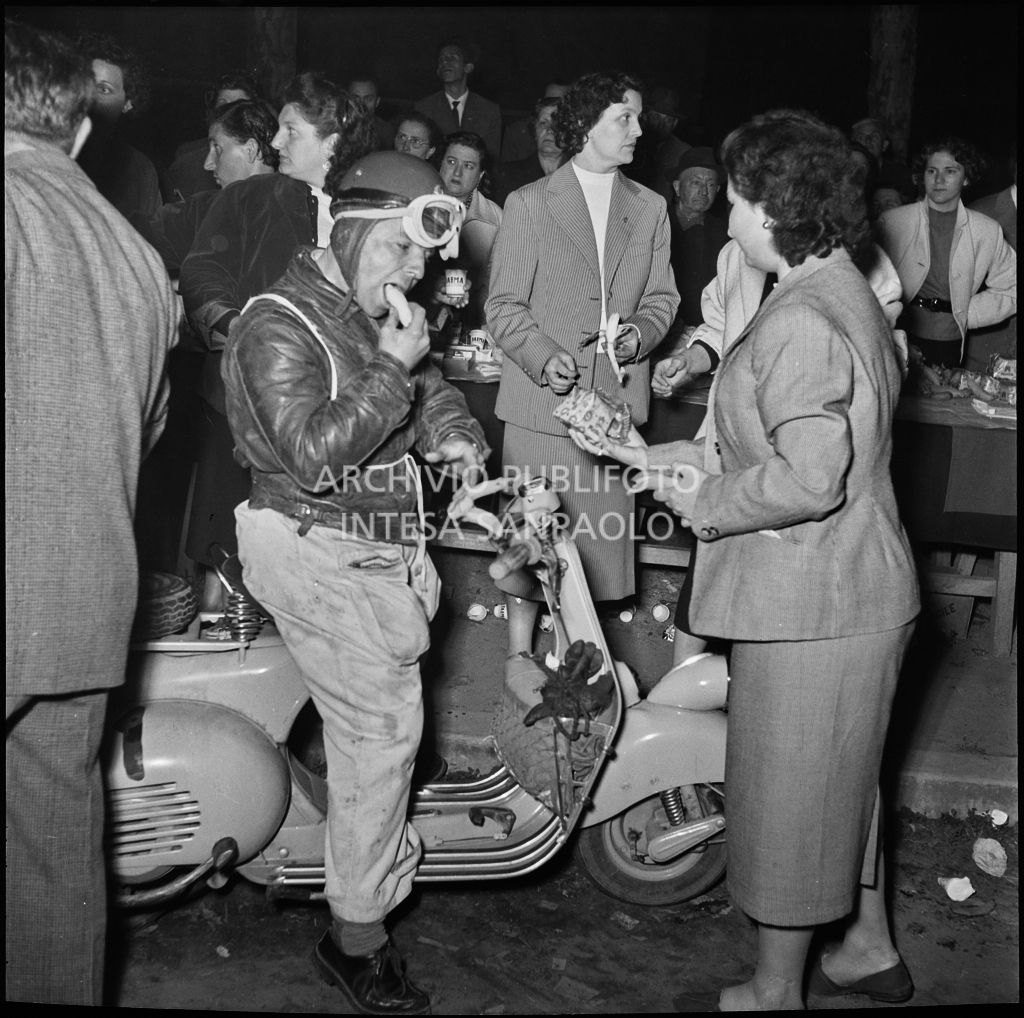 Un concorrente mangia una banana in sella alla sua Vespa durante una pausa dalla competizione sportiva 1000 Km Vespistica del 1954<br>216445