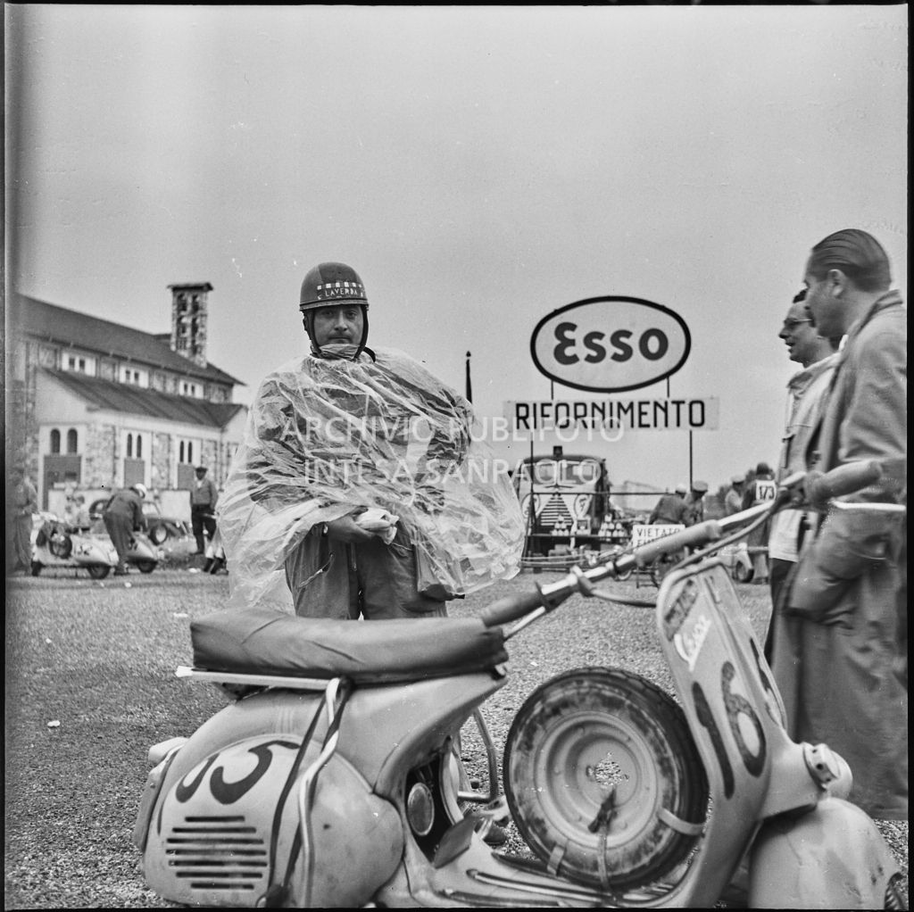 Un concorrente (numero di gara 165) con un impermeabile in un momento di pausa dalla competizione sportiva 1000 Km Vespistica del 1954<br>216467
