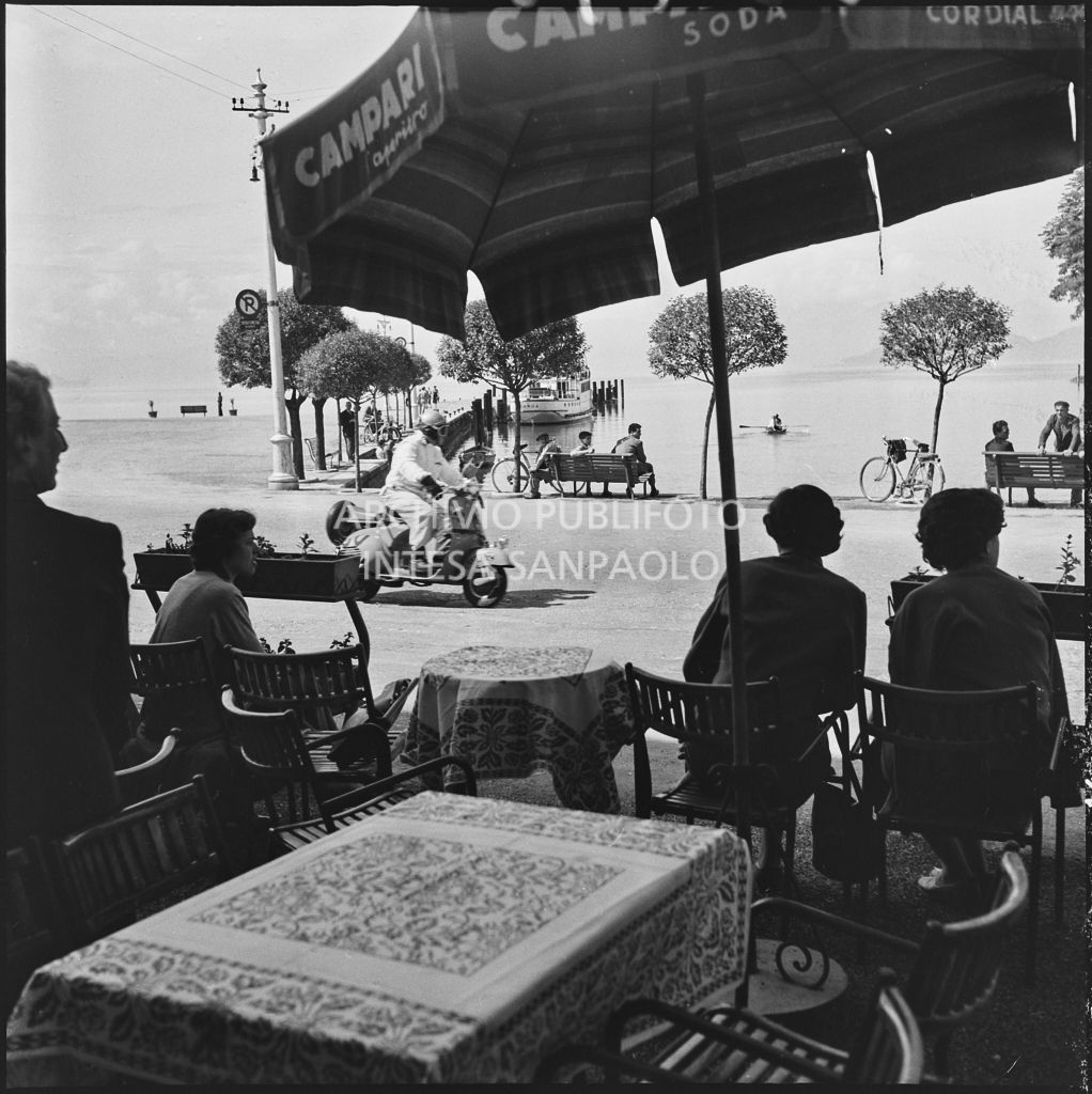Alcuni avventori di un bar osservano il passaggio di un concorrente della competizione sportiva 1000 Km Vespistica del 1954 a Peschiera del Garda<br>216475