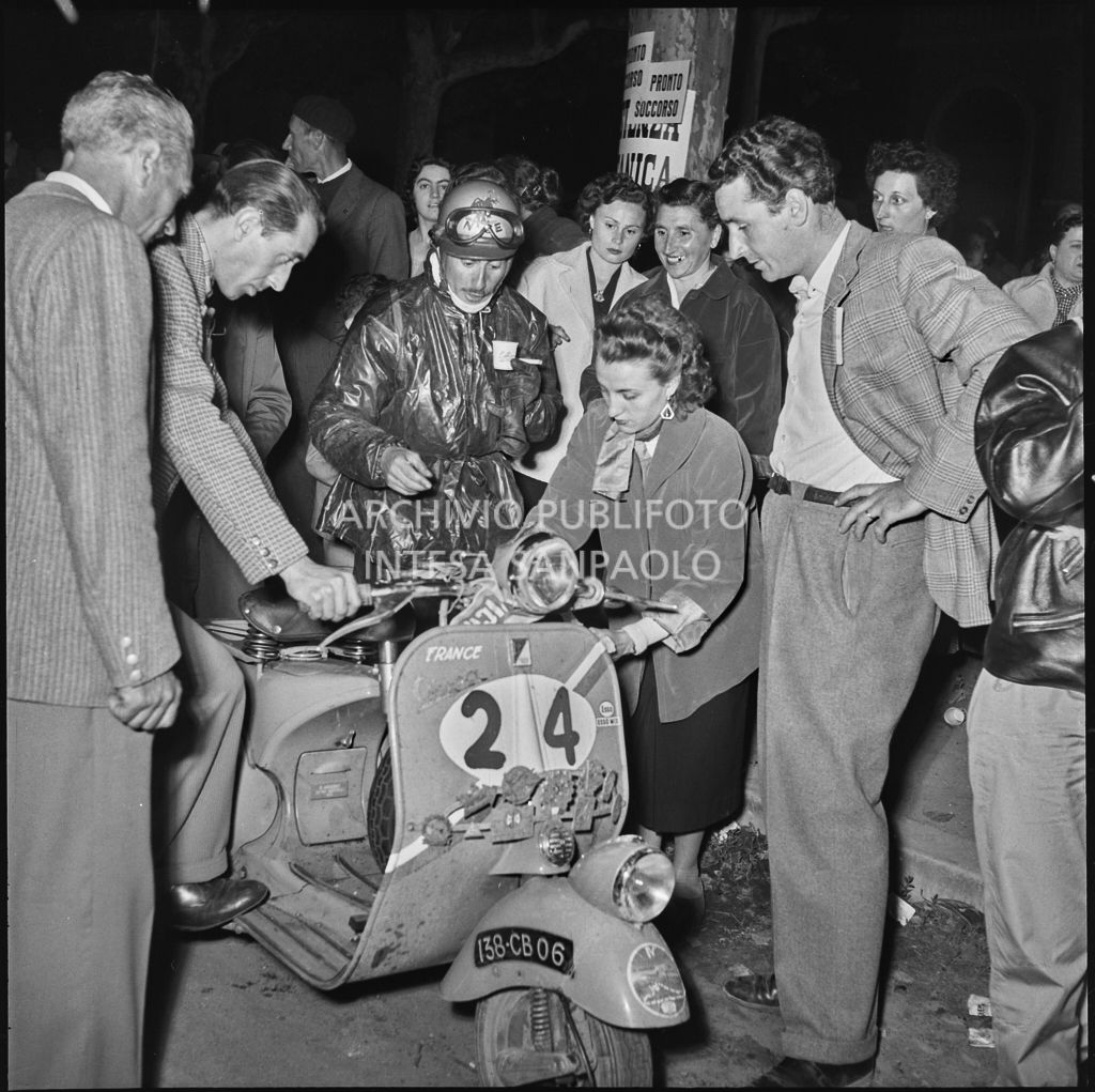 Il concorrente Rudy Aragno del Vespa Club di Parigi (numero di gara 24), circondato da lacune persone, beve un caffè vicino al suo scooter in un momento di pausa dalla competizione sportiva 1000 Km Vespistica del 1954<br>216489