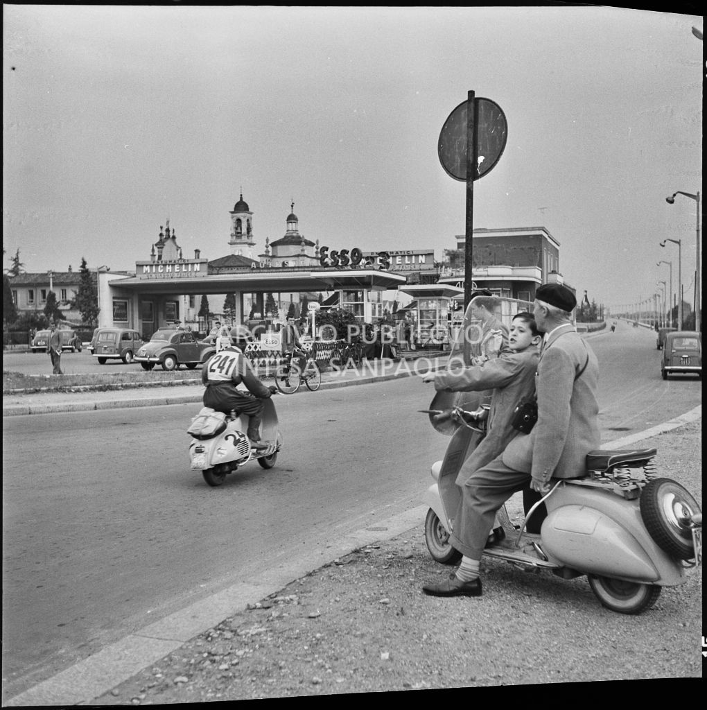 Alcuni spettatori, a bordo di una Vespa, osservano il passaggio di un concorrente (numero di gara 241) della competizione sportiva 1000 Km Vespistica del 1954 nei pressi della Certosa di Milano<br>216513
