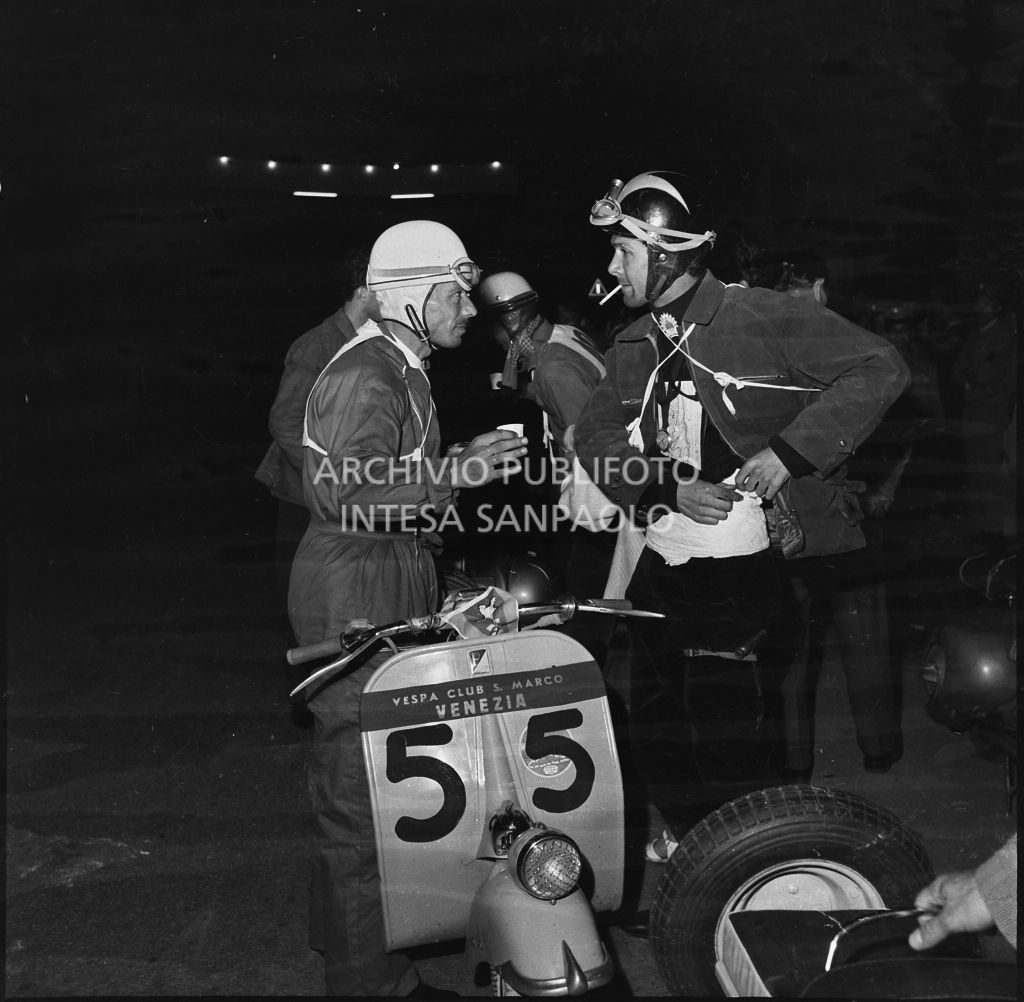 Il concorrente Elia Morchietti del Vespa Club di Venezia (numero di gara 55) conversa con un altro concorrente durante una pausa dalla competizione sportiva 1000 Km Vespistica del 1954<br>216526