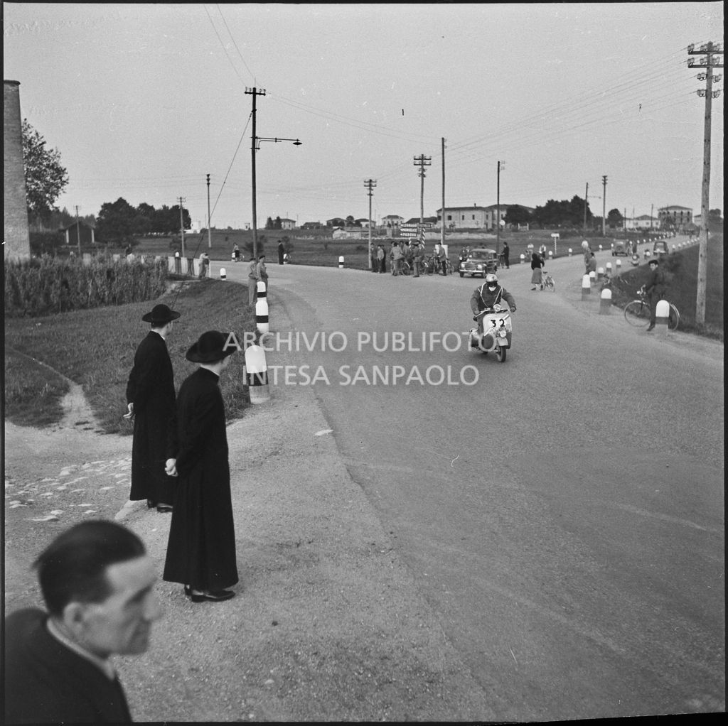 Alcuni preti osservano il passaggio di un concorrente (numero di gara 32) durante la competizione sportiva 1000 Km Vespistica del 1954<br>216534